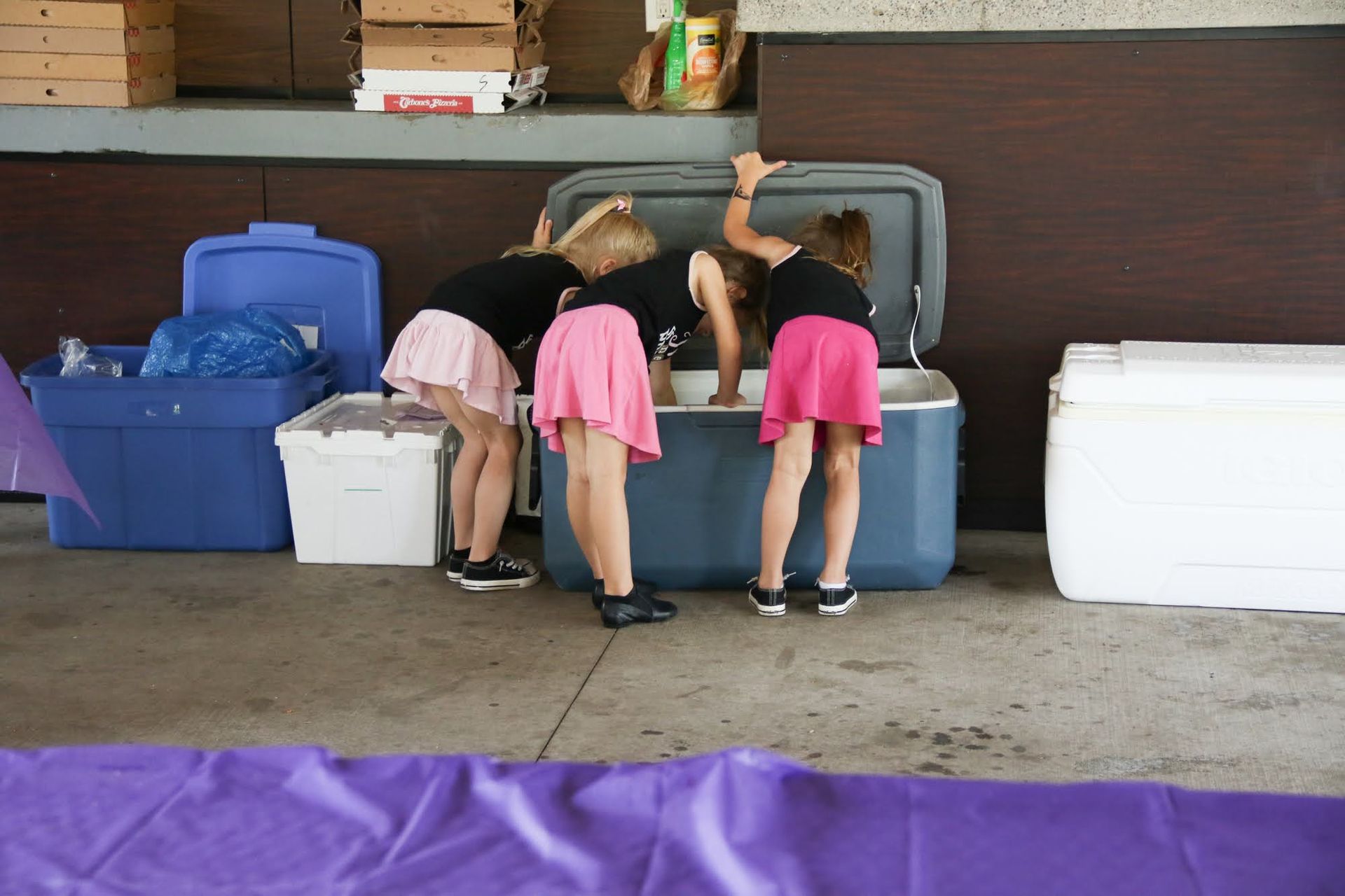 Three people in pink skirts and black shirts look into a large cooler.