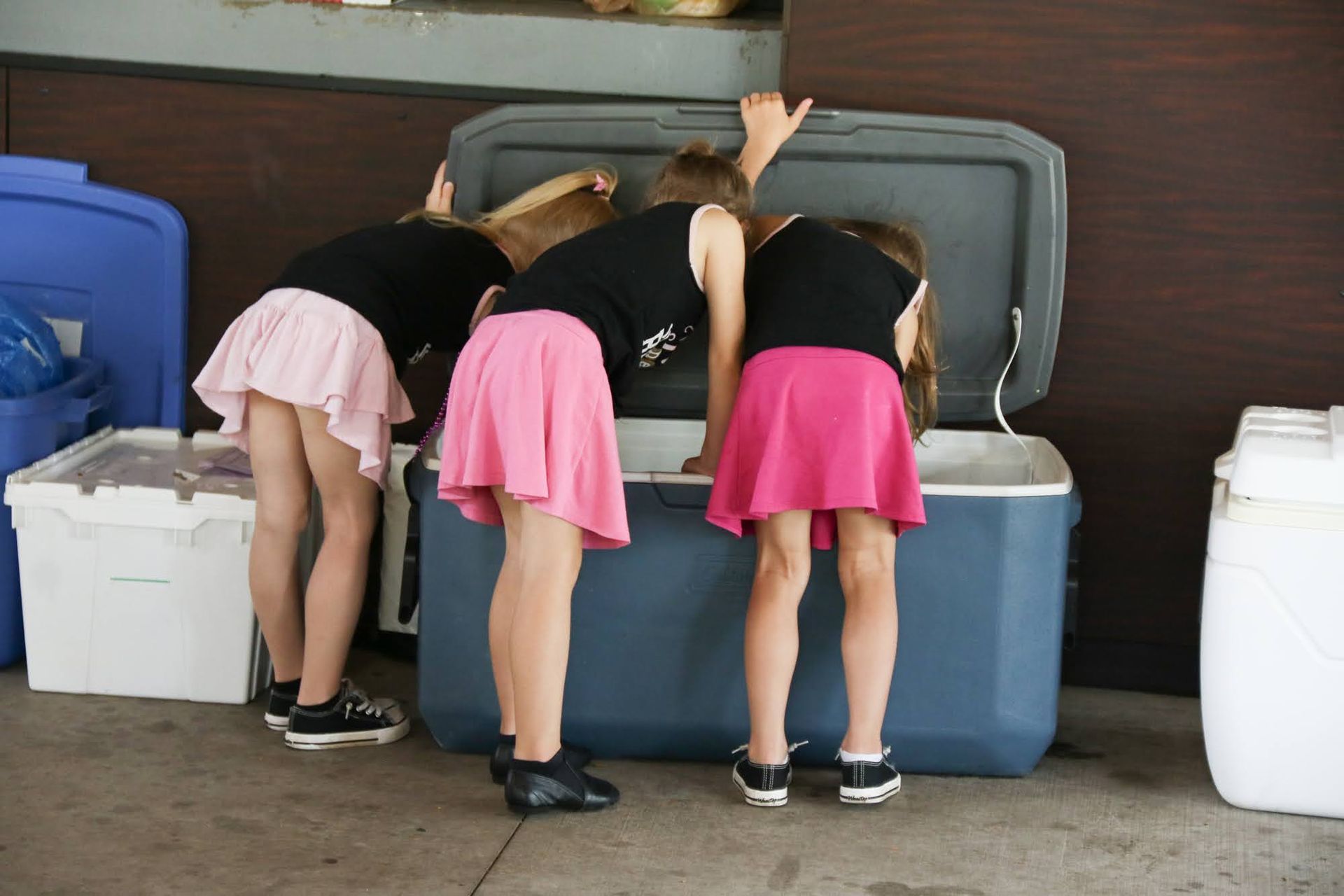 Three children look inside a large blue cooler. One child wears a pink skirt, two wear black tops.