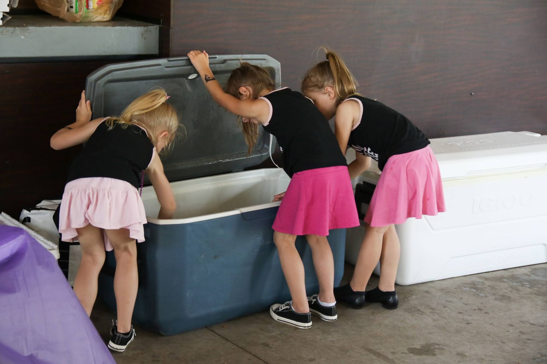 Three people, wearing pink skirts and black tops, look inside a blue cooler.