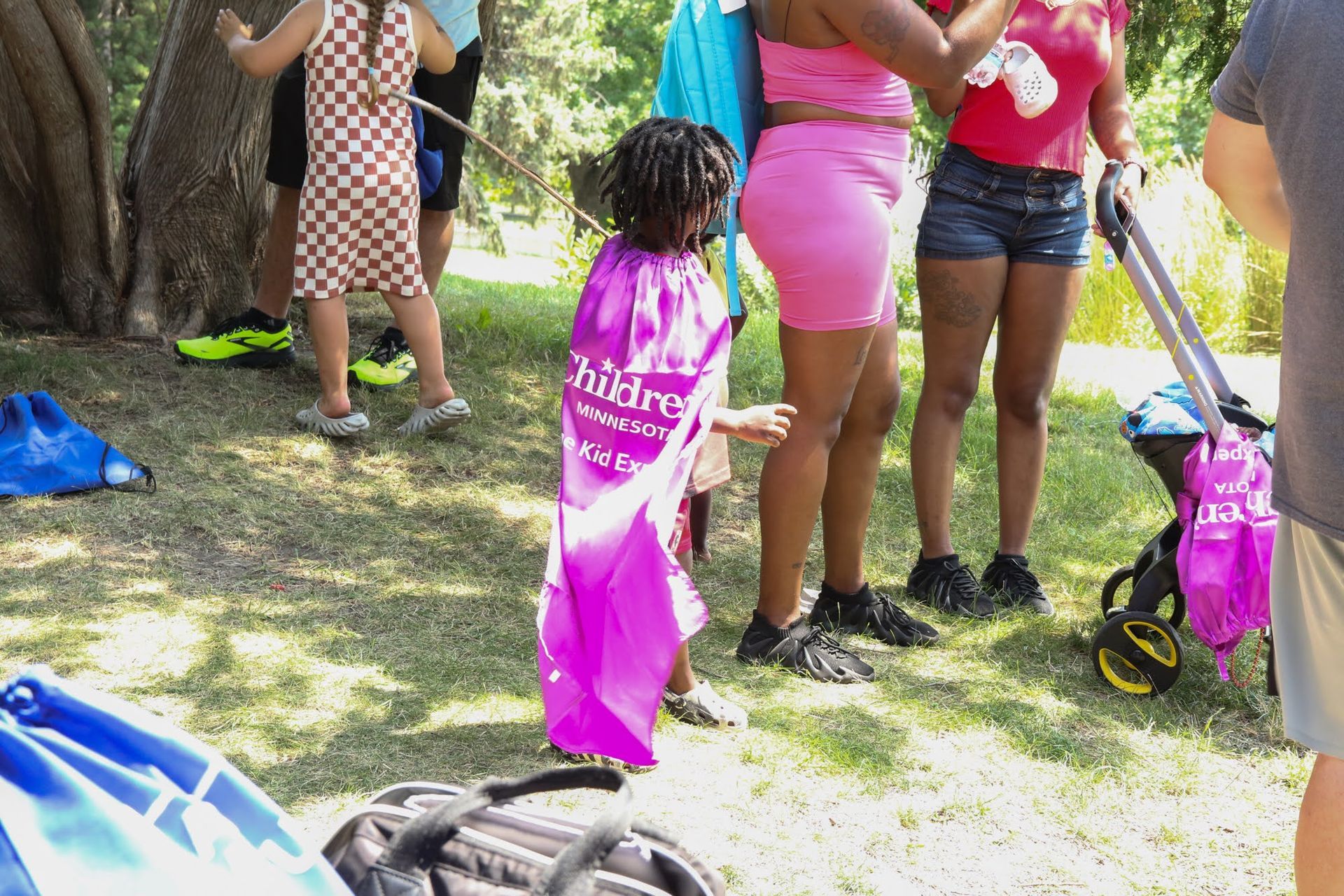 Child in purple cape stands on grass with others, near a stroller.