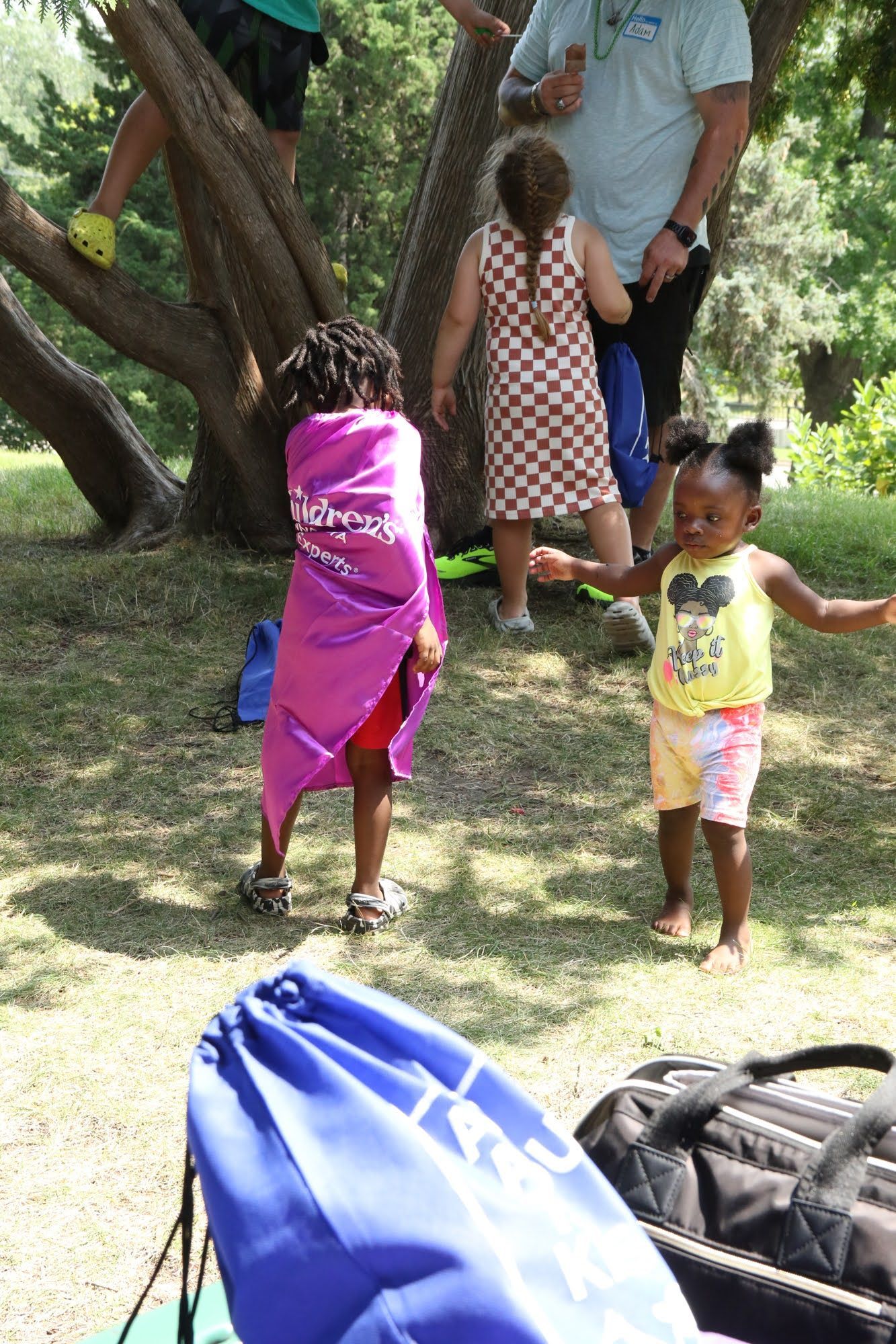Children playing in a park, one wrapped in pink. A person is climbing a tree. Blue bag in the foreground.