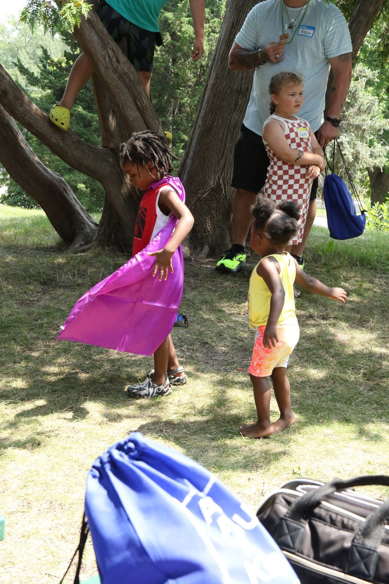 Two children playing outdoors, one in pink, one in yellow. People in the background by a tree. Blue bag in the foreground.