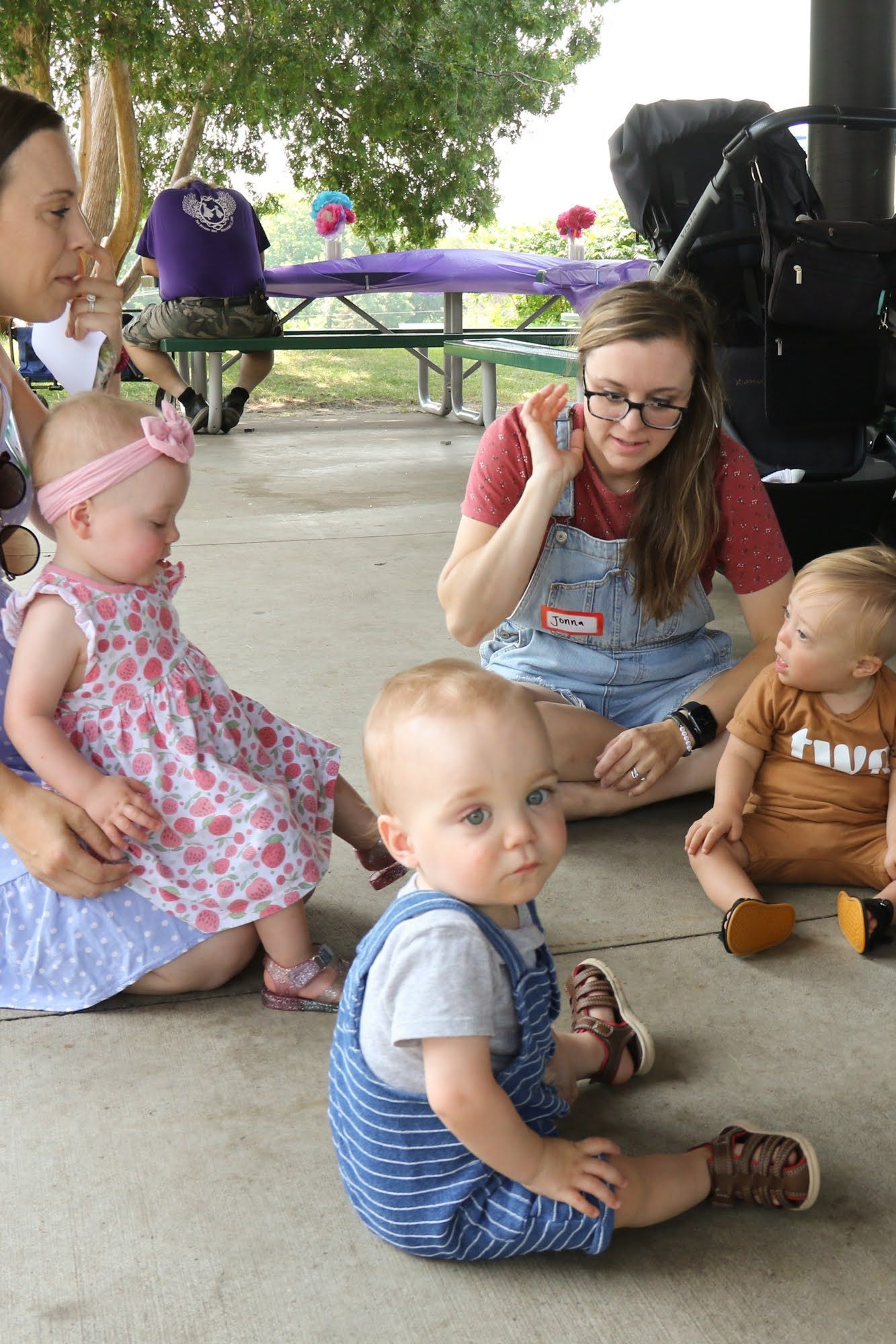 Babies with two women in a park. Babies in the foreground. One baby in blue overalls. Woman in overalls with a baby on the right.