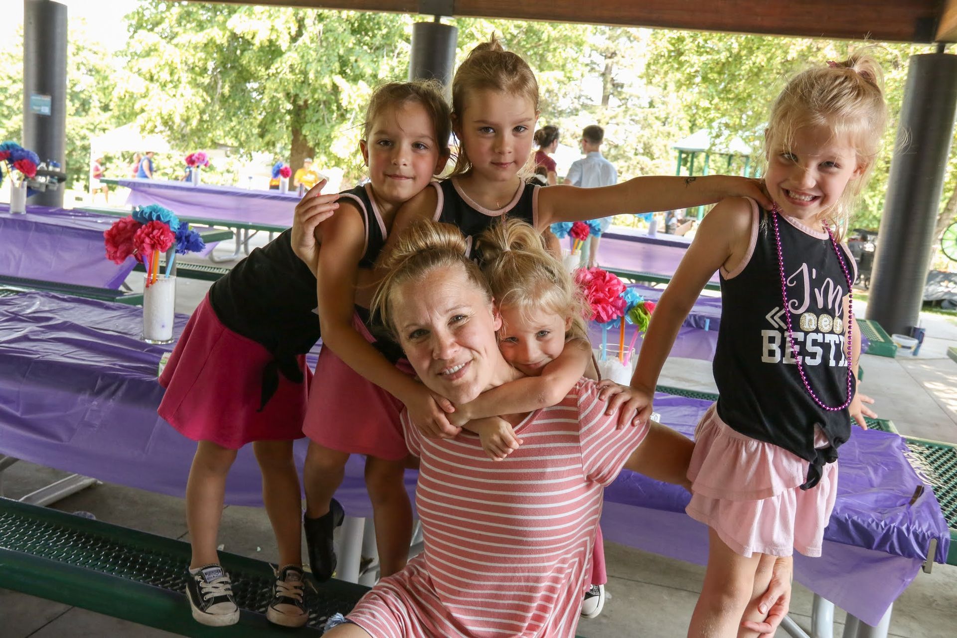 Woman and four young girls smiling, hugging, at a park with decorated tables.