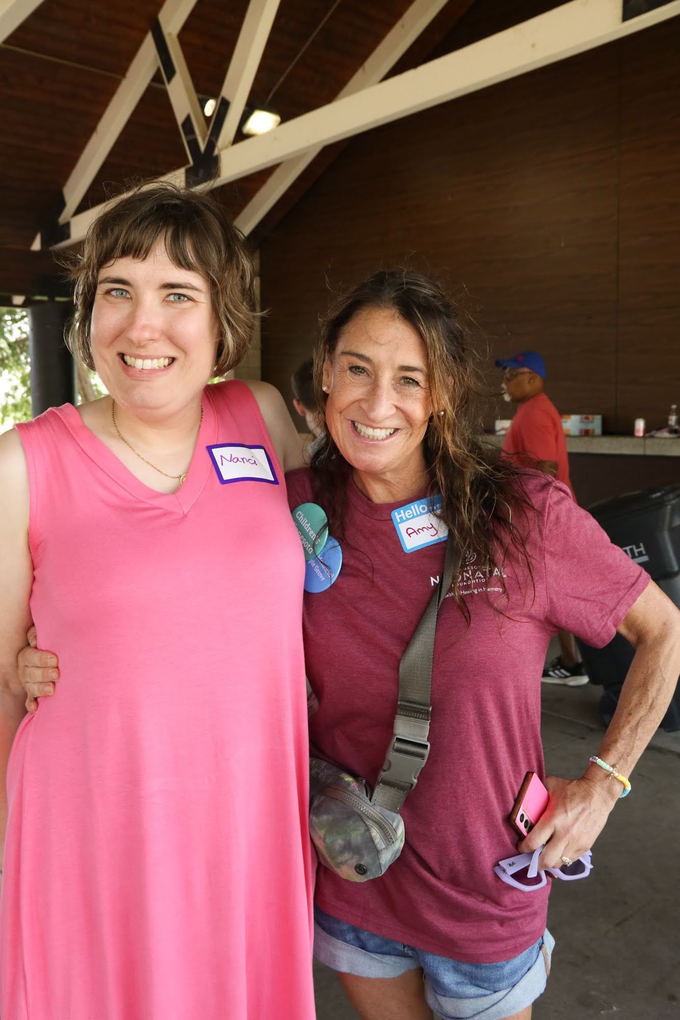 Two women smiling, arm in arm, under a pavilion. One wears a pink dress, the other a maroon shirt and denim shorts.