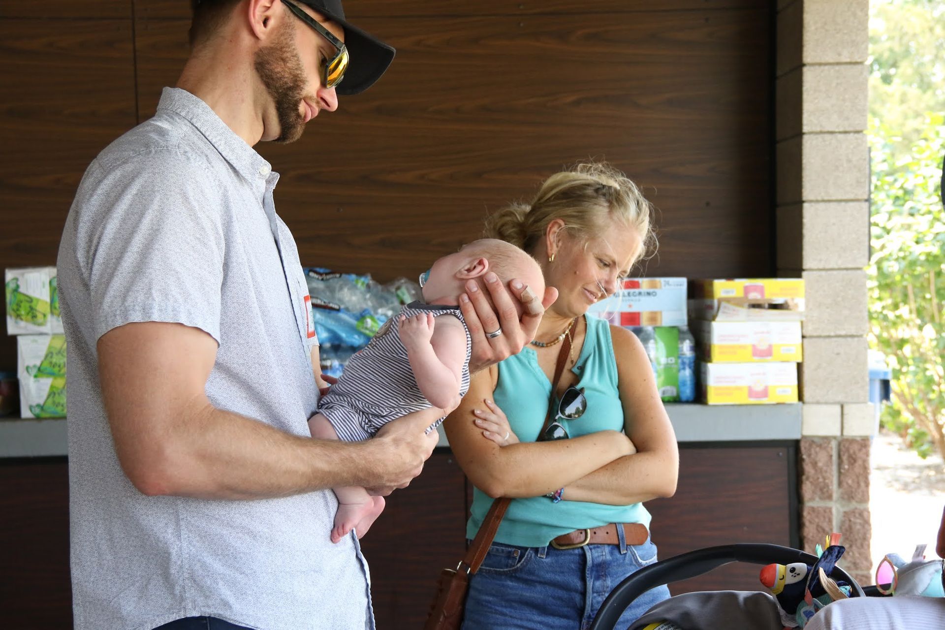 Man holding baby, woman watches, standing near a stroller. Outdoors with brown and tan structures.