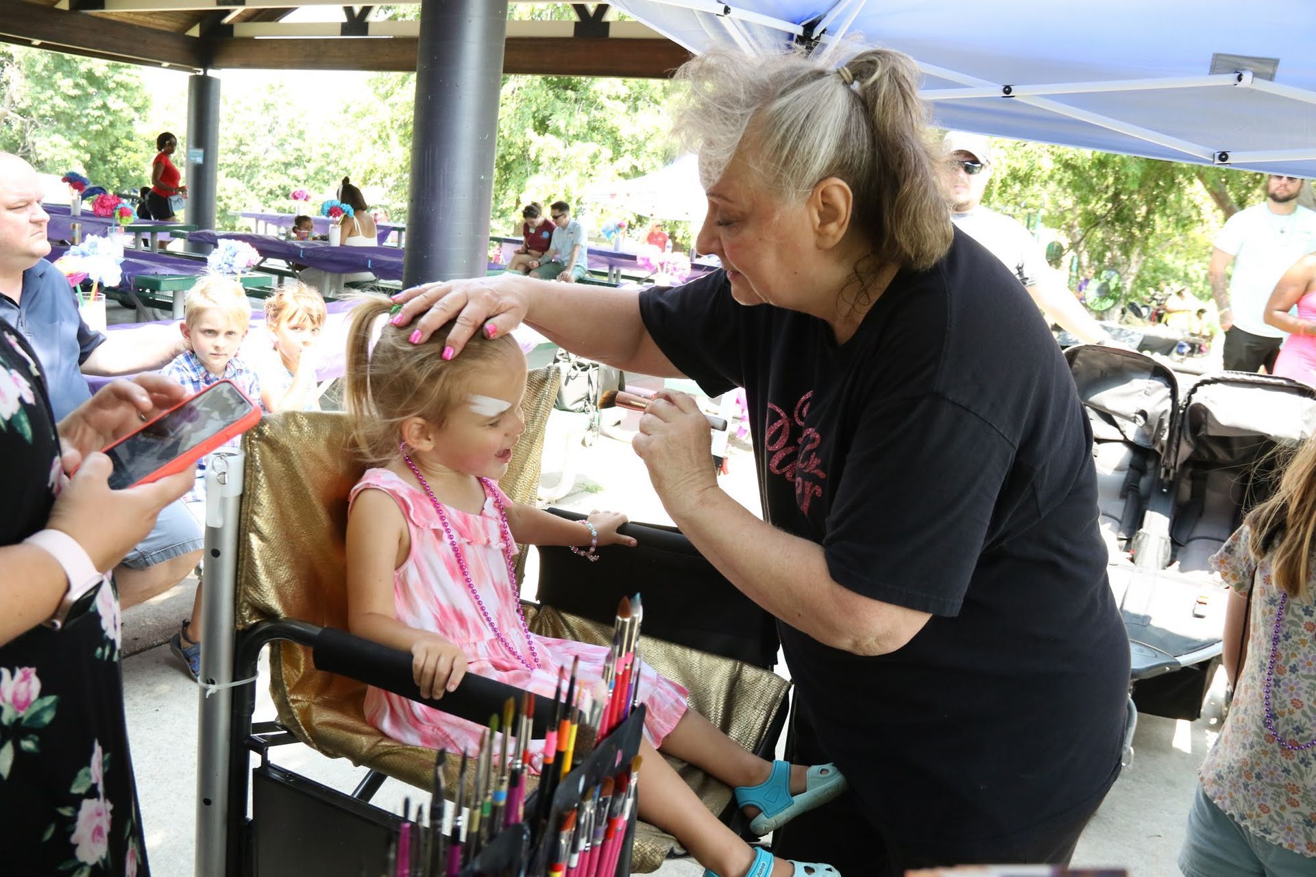 Woman paints face of a child in outdoor setting, with other children and adults present.