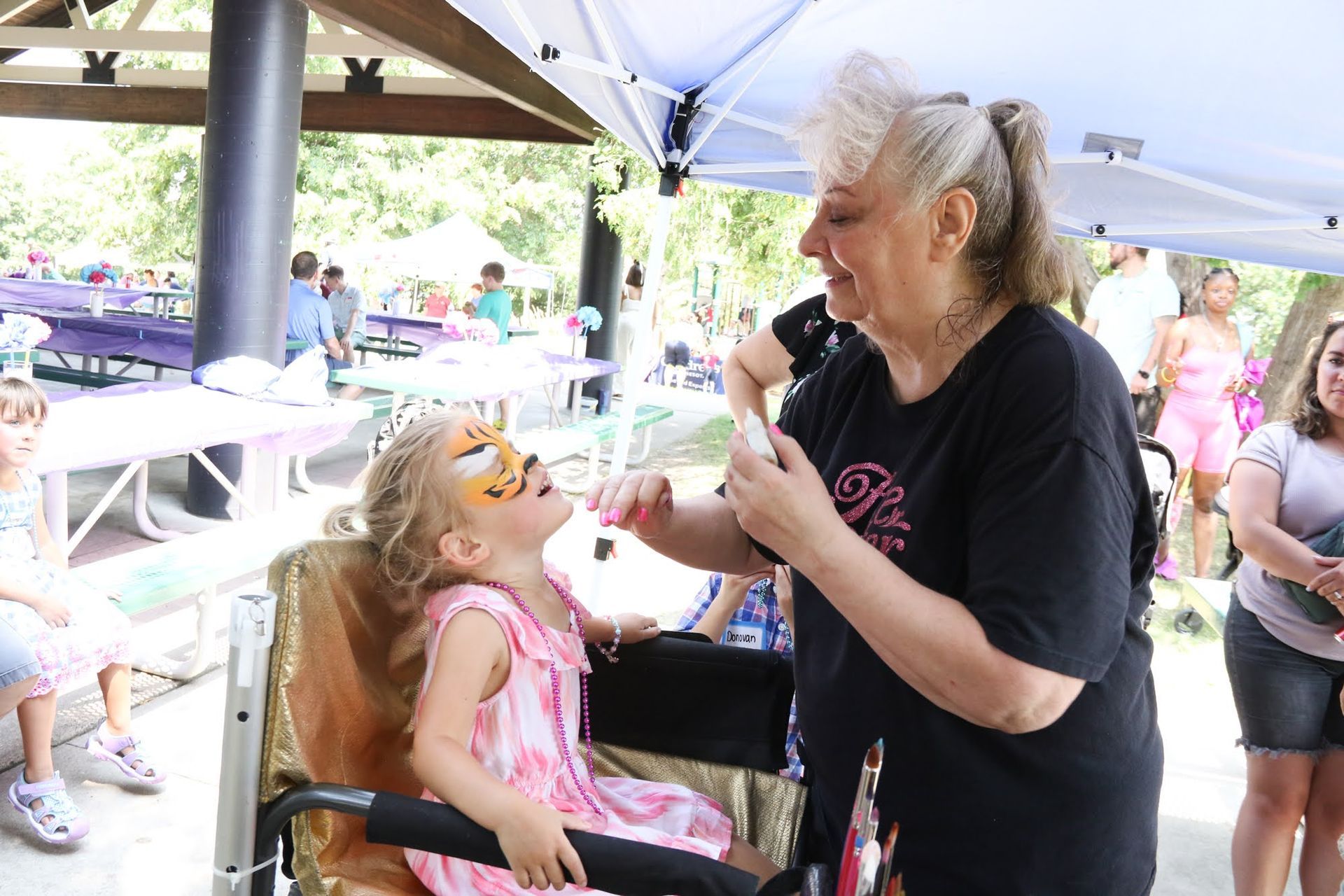 Woman painting a tiger face on a child's face at an outdoor event.