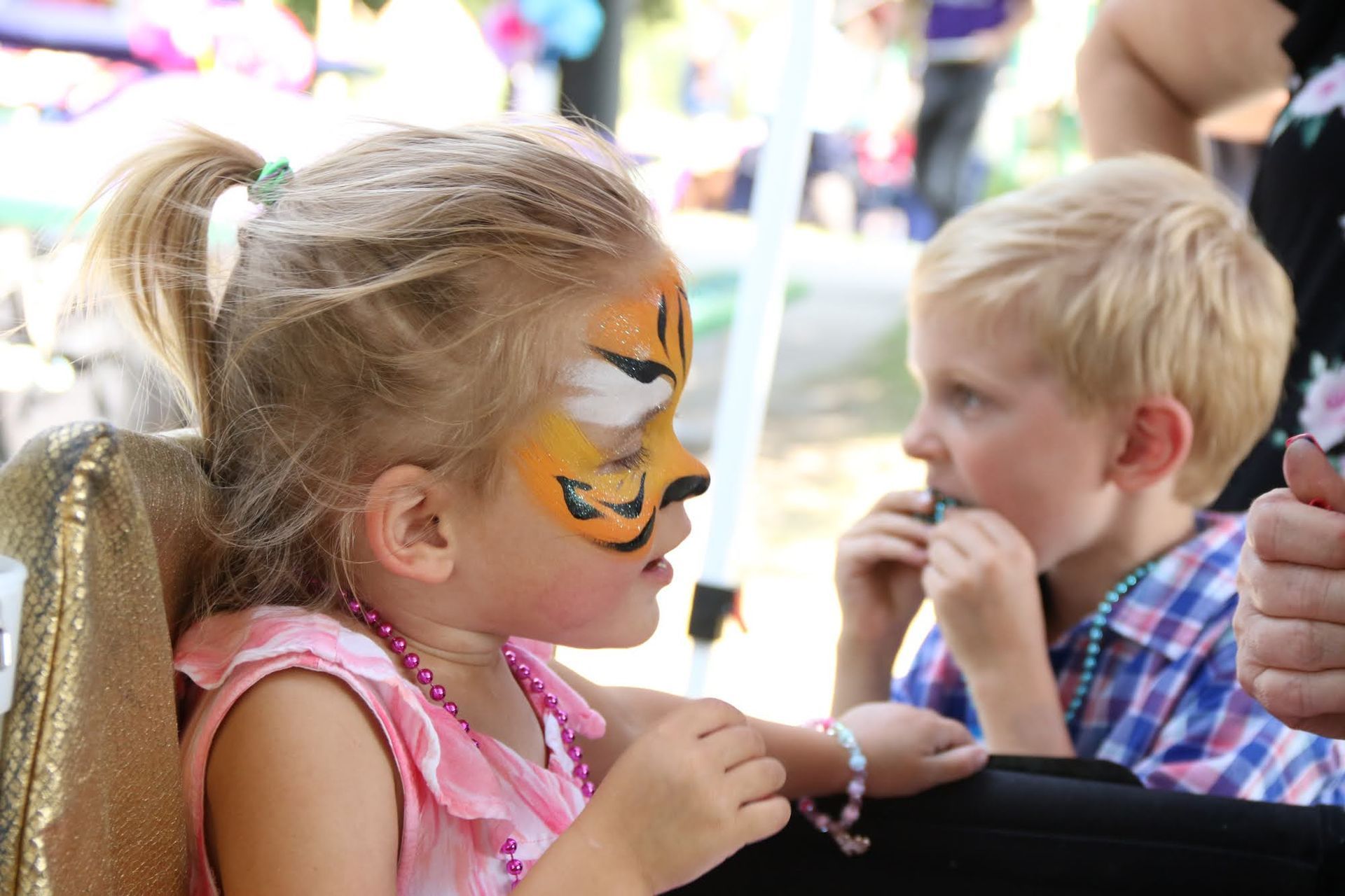 Girl with tiger face paint looks at a boy eating something outdoors.
