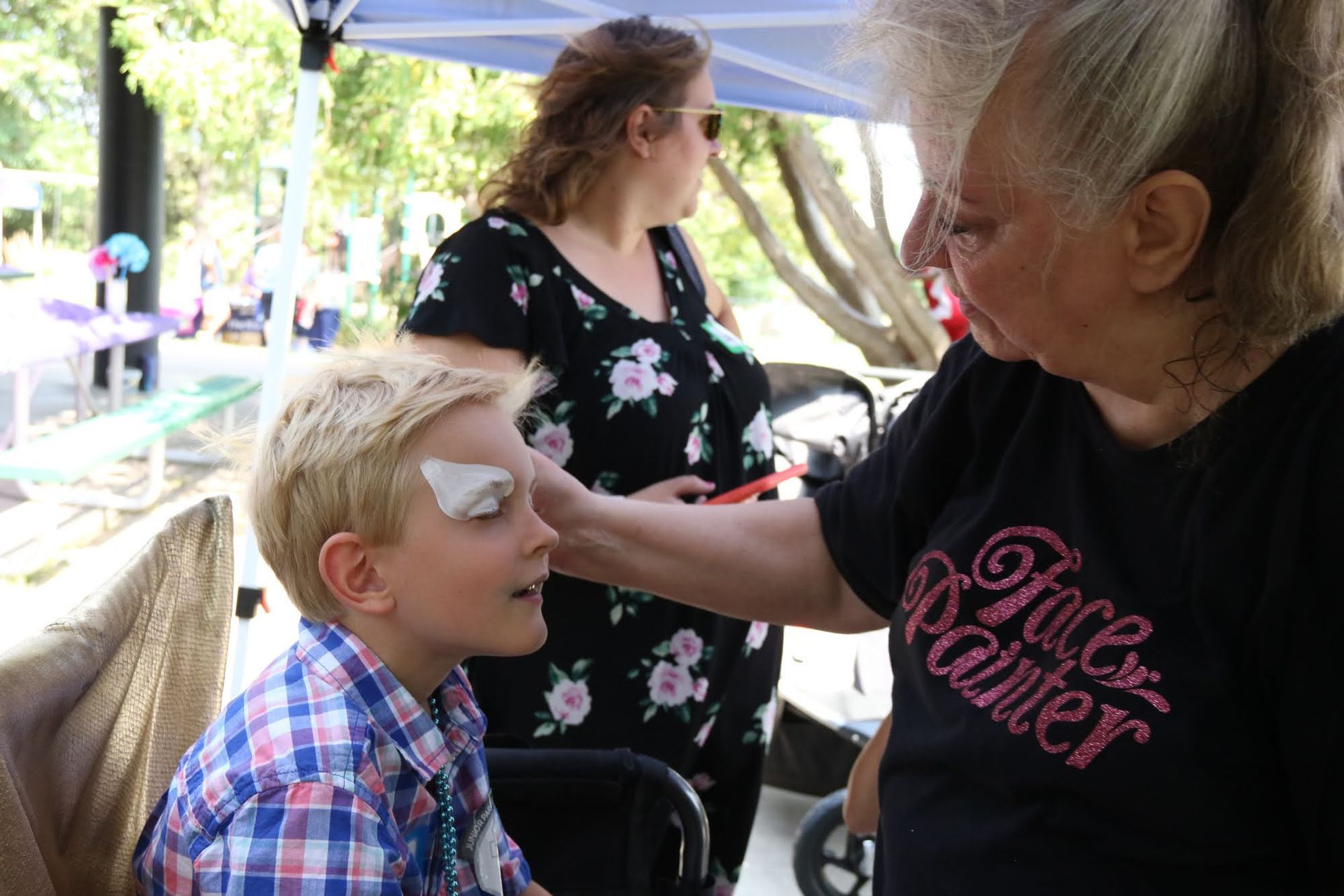 Person with face paint being applied to their eye at an outdoor event. Another person and a stroller are in the background.