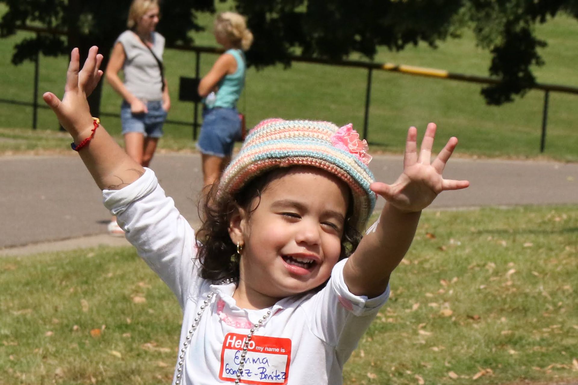 Young child with arms raised, smiling, wearing a hat and name tag, outdoors on a sunny day.