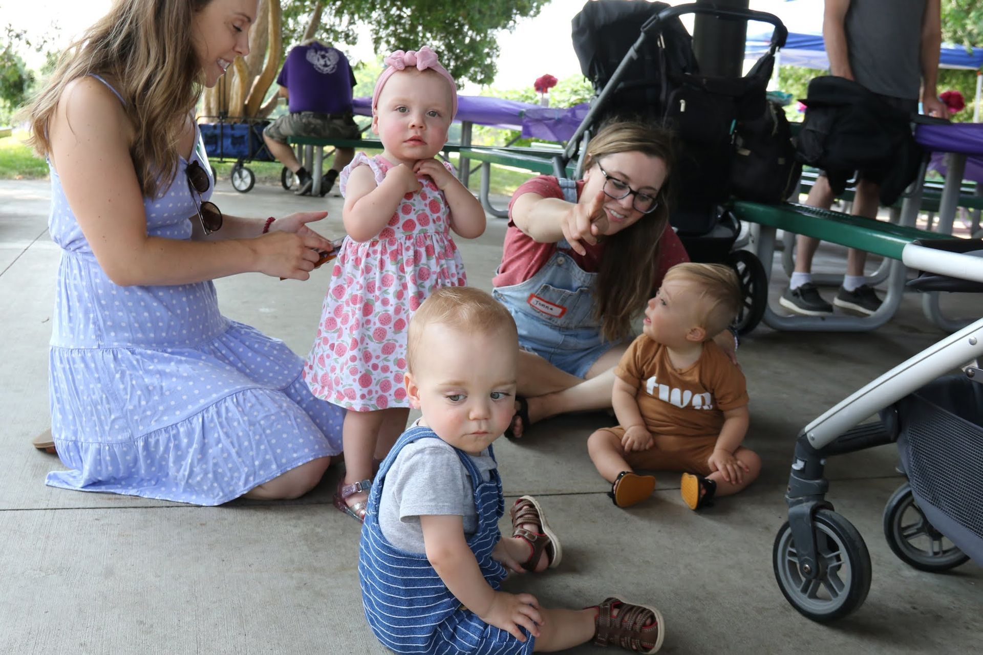 Four babies and two women gathered outdoors near strollers and picnic tables.