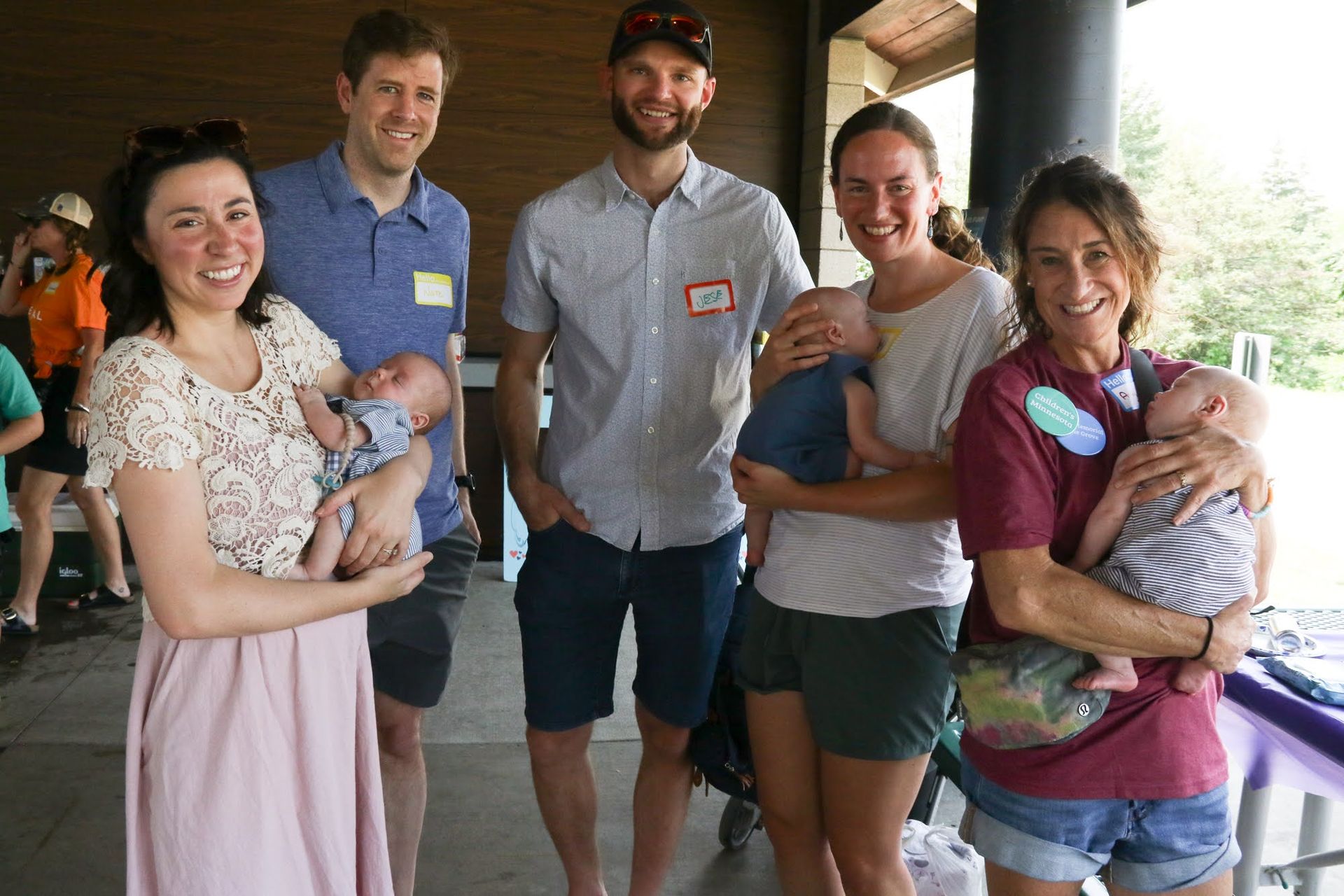 Group of adults holding babies, smiling outdoors under a shelter.