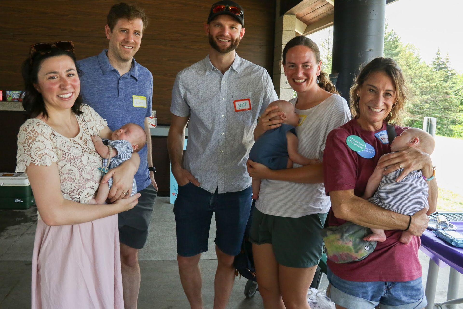 Six people at a park, holding babies. Two women and three men smiling, sunny day.