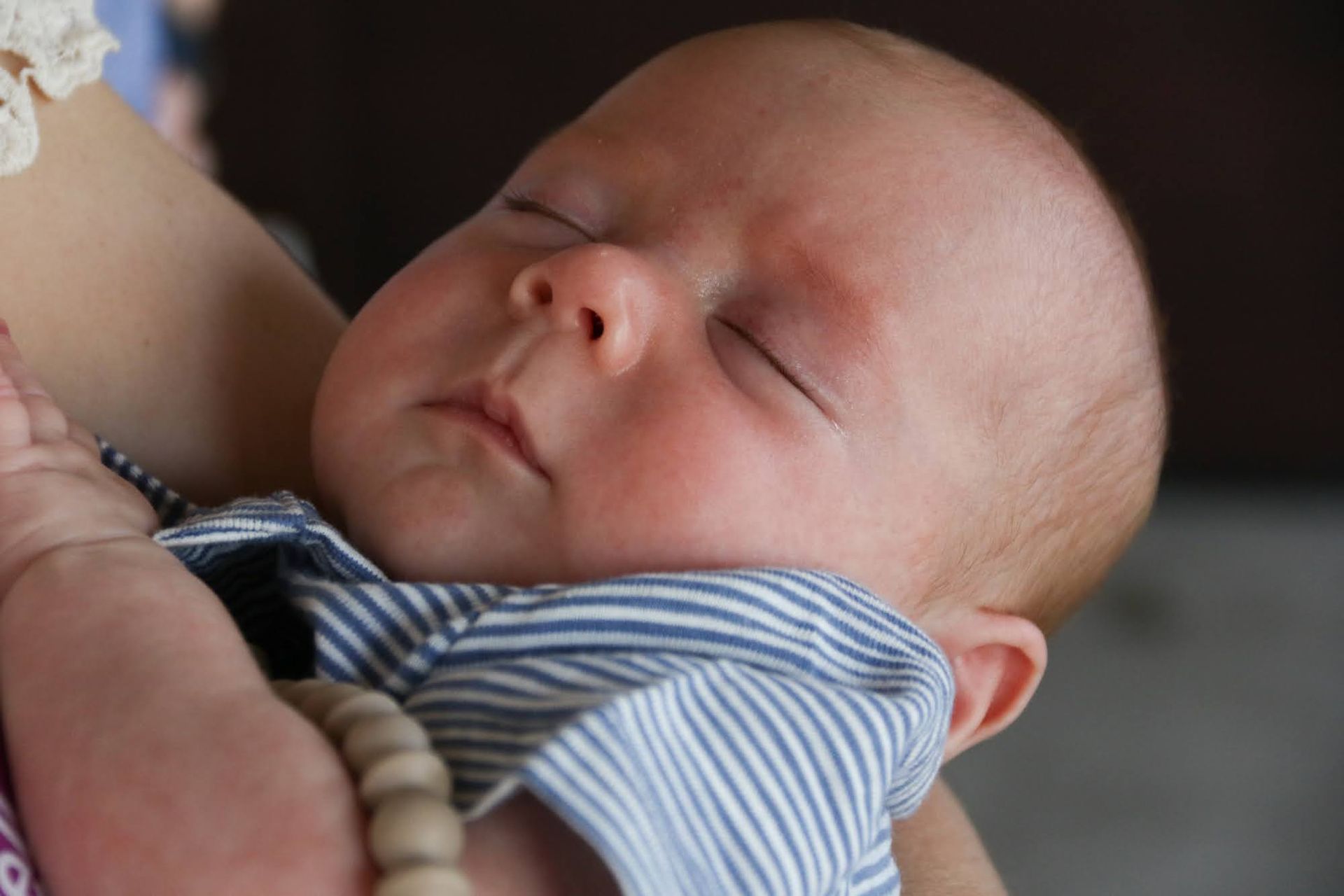 Sleeping baby held in arms, wearing blue and white striped shirt, wooden beads.