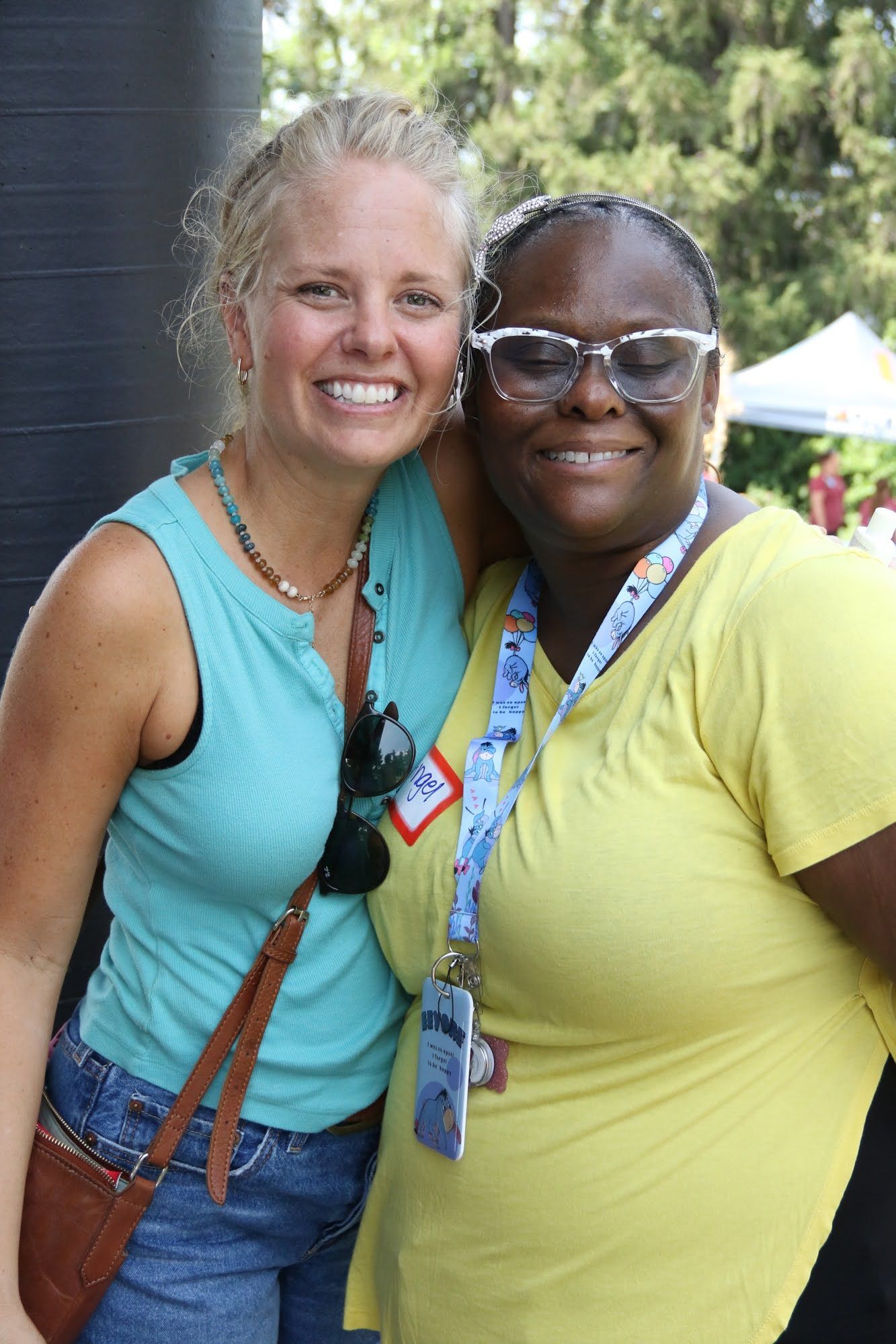 Two women smiling, posing for a photo. One in a blue shirt, the other in a yellow shirt with glasses. Outdoor setting.