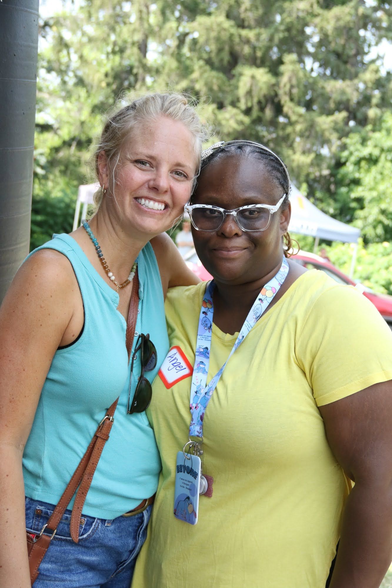 Two women smile, arms around each other outdoors; one in turquoise top, the other in yellow.