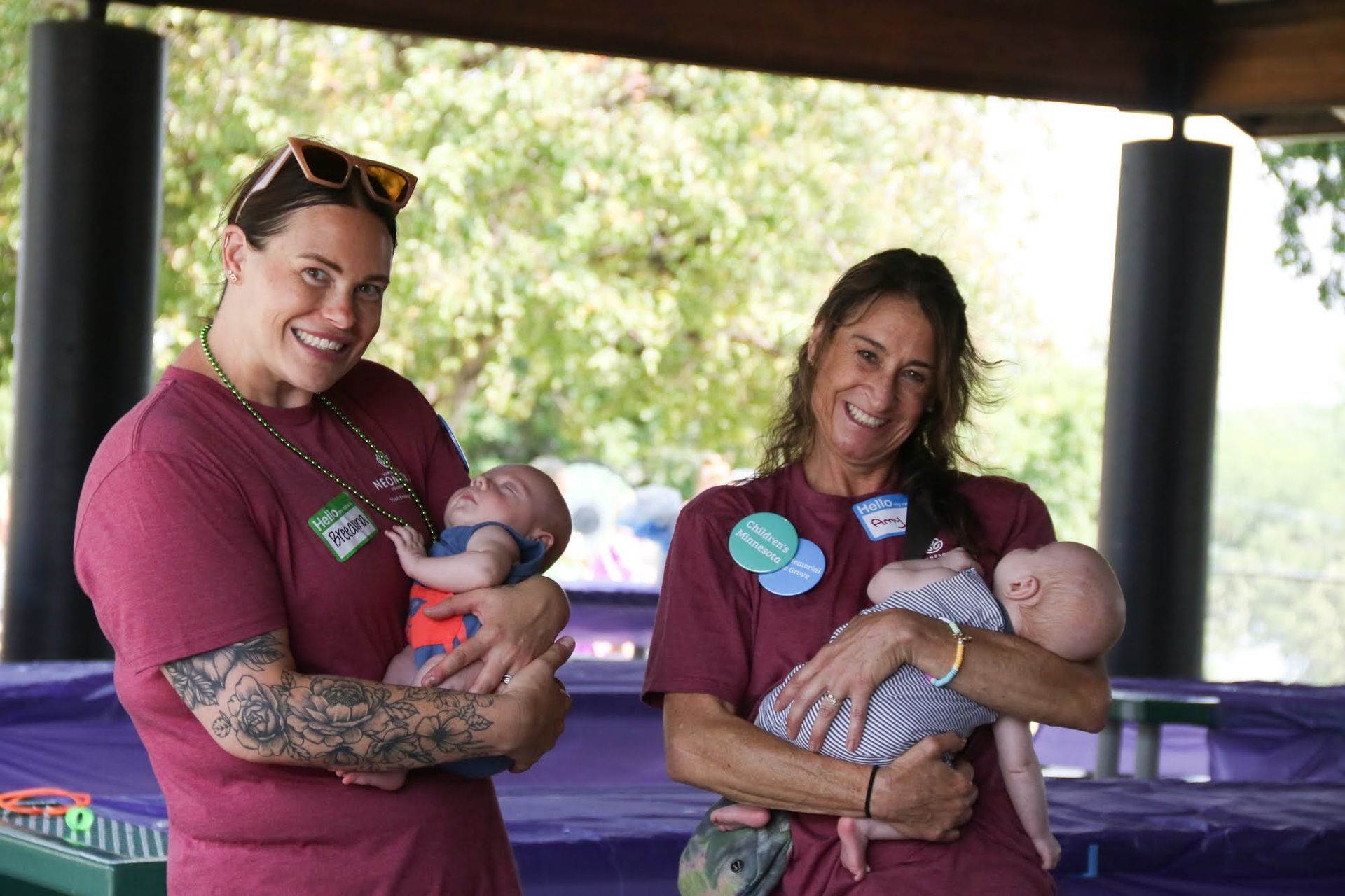 Two people holding babies, smiling outdoors under a shelter. They wear maroon shirts and name tags.