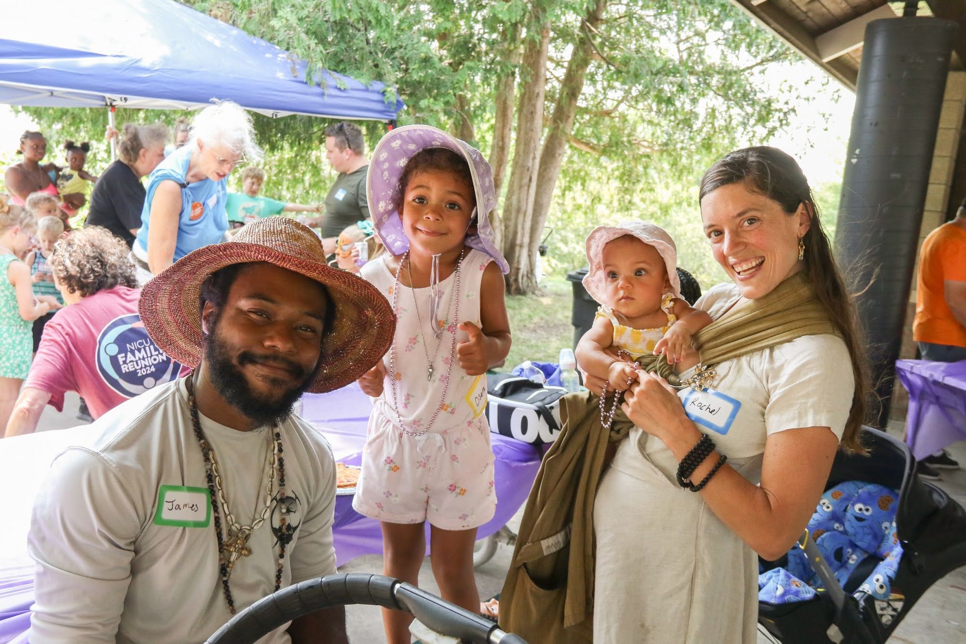 Group of people at an outdoor event smiling. Man with hat, woman holding baby, child in hat.