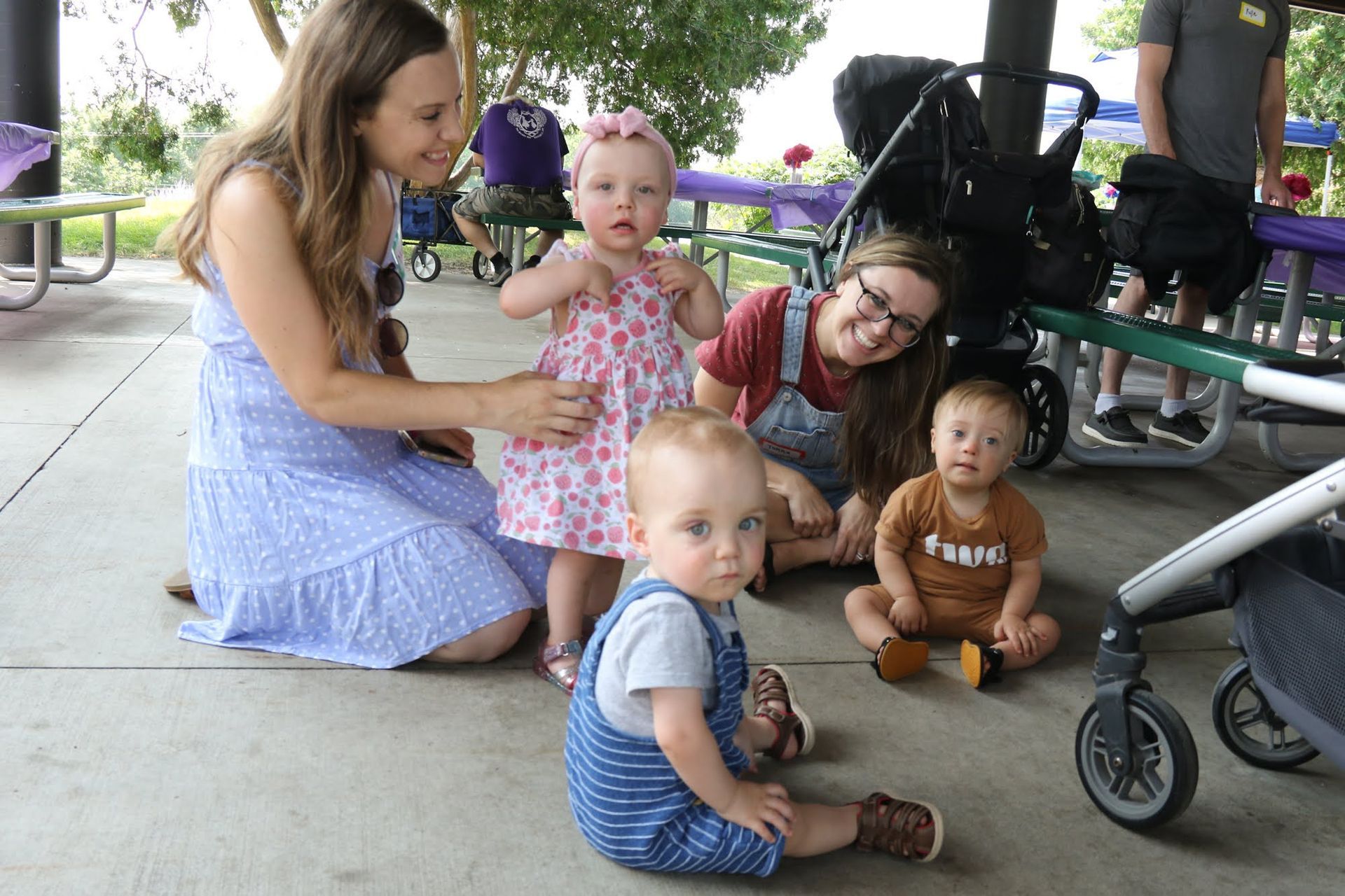 Four babies and two adults at a park. Babies sit on the ground, adults smile nearby.
