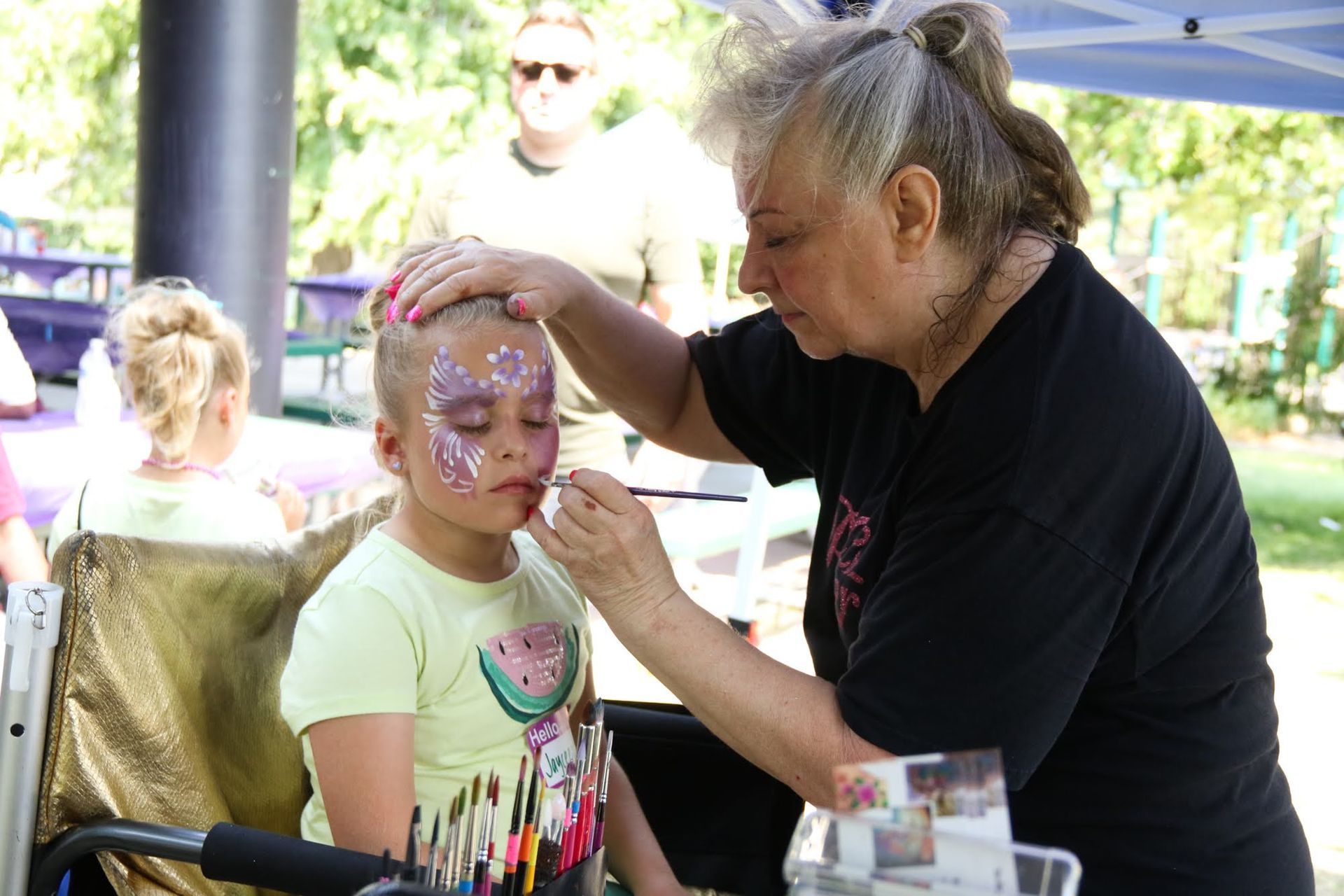 Woman paints a design on a child's face with glitter and pink paint, outside under a tent.