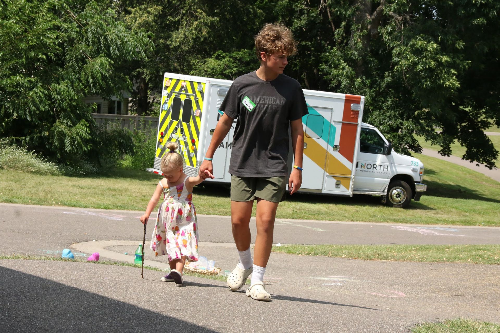 A young person walks with a small child on a path, ambulance in the background.