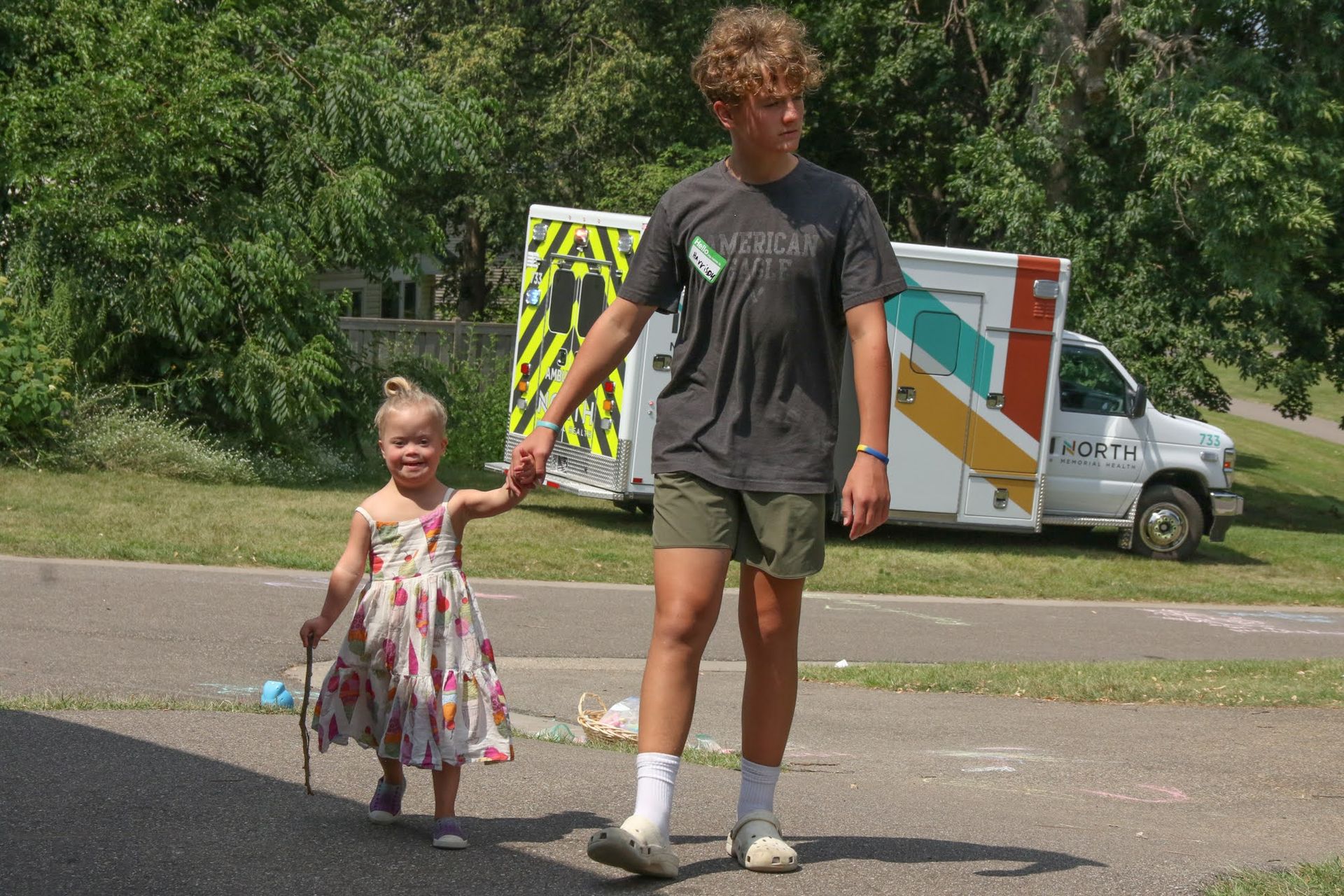 A young person holds hands with a child while walking on a road with an ambulance in the background.