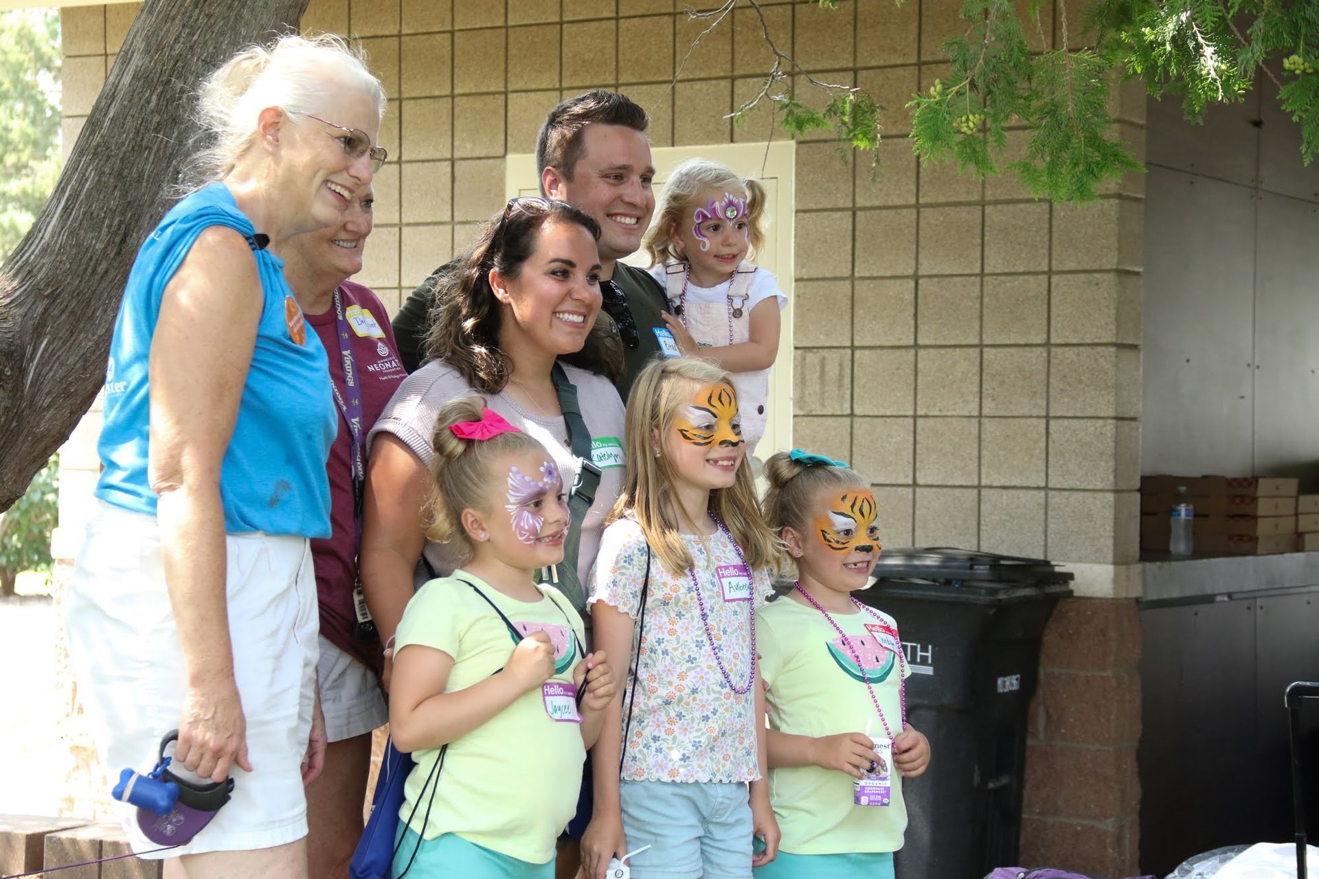 Group smiling for a photo; people with face paint, in front of a building with a tree.
