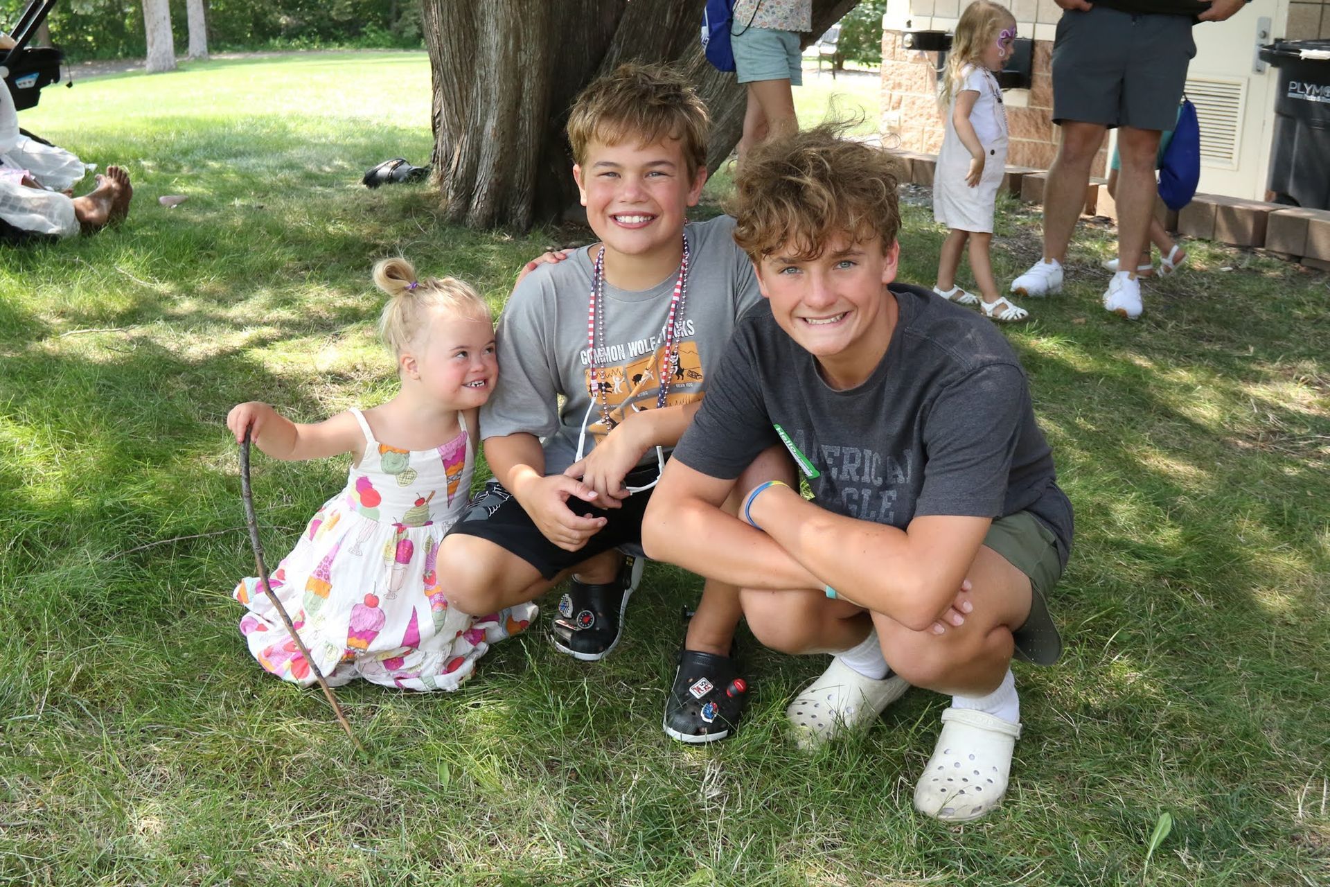 Three children squatting on grass, smiling. A little girl holds a string, two boys flank her.
