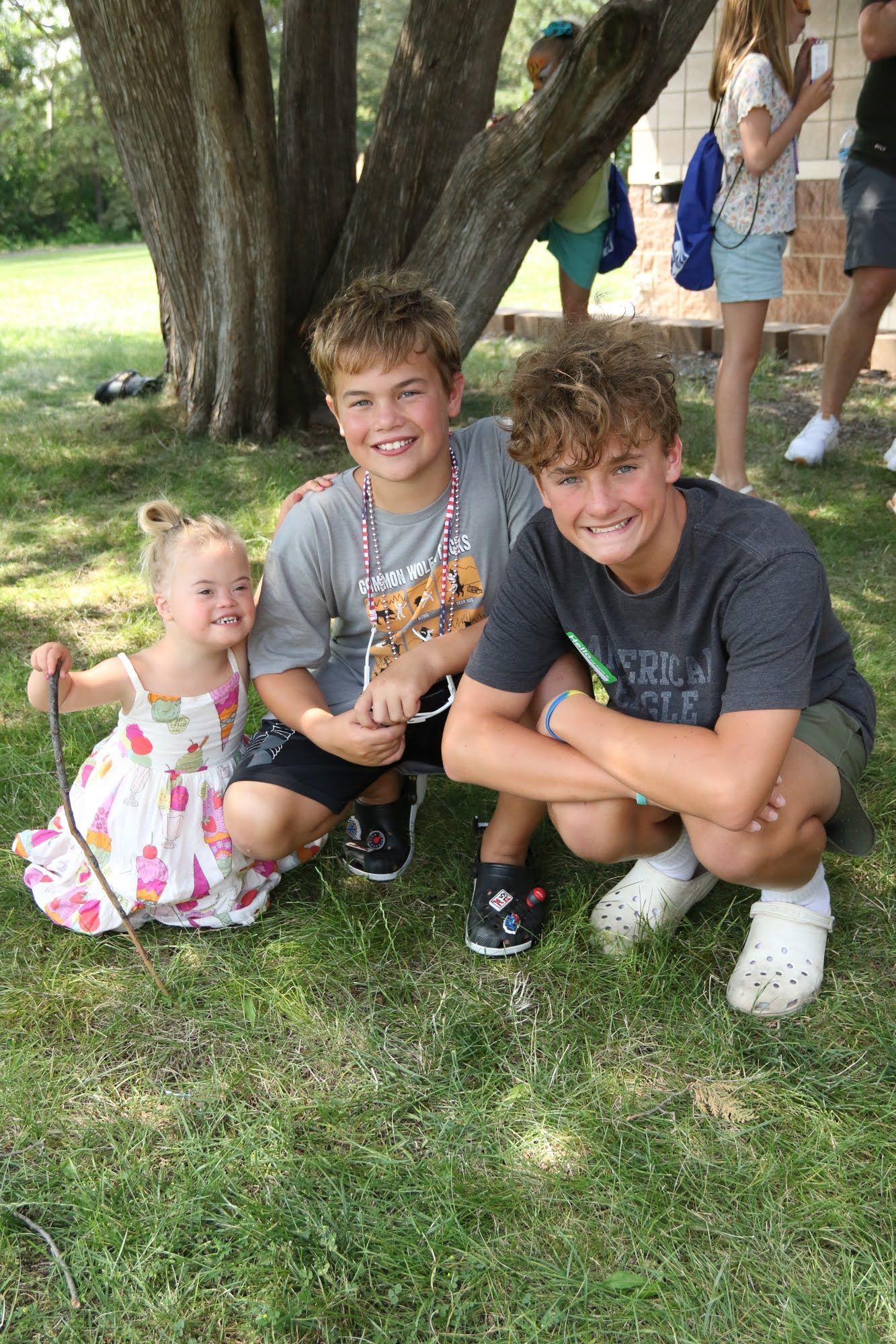 Three smiling children in a grassy area, one kneeling in a floral dress, two squatting in shorts and shirts.