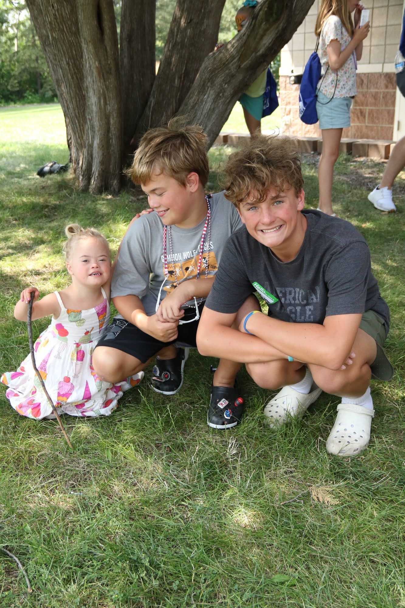 Three smiling children crouching on grass near a tree; one wears a floral dress, two wear t-shirts and shorts.