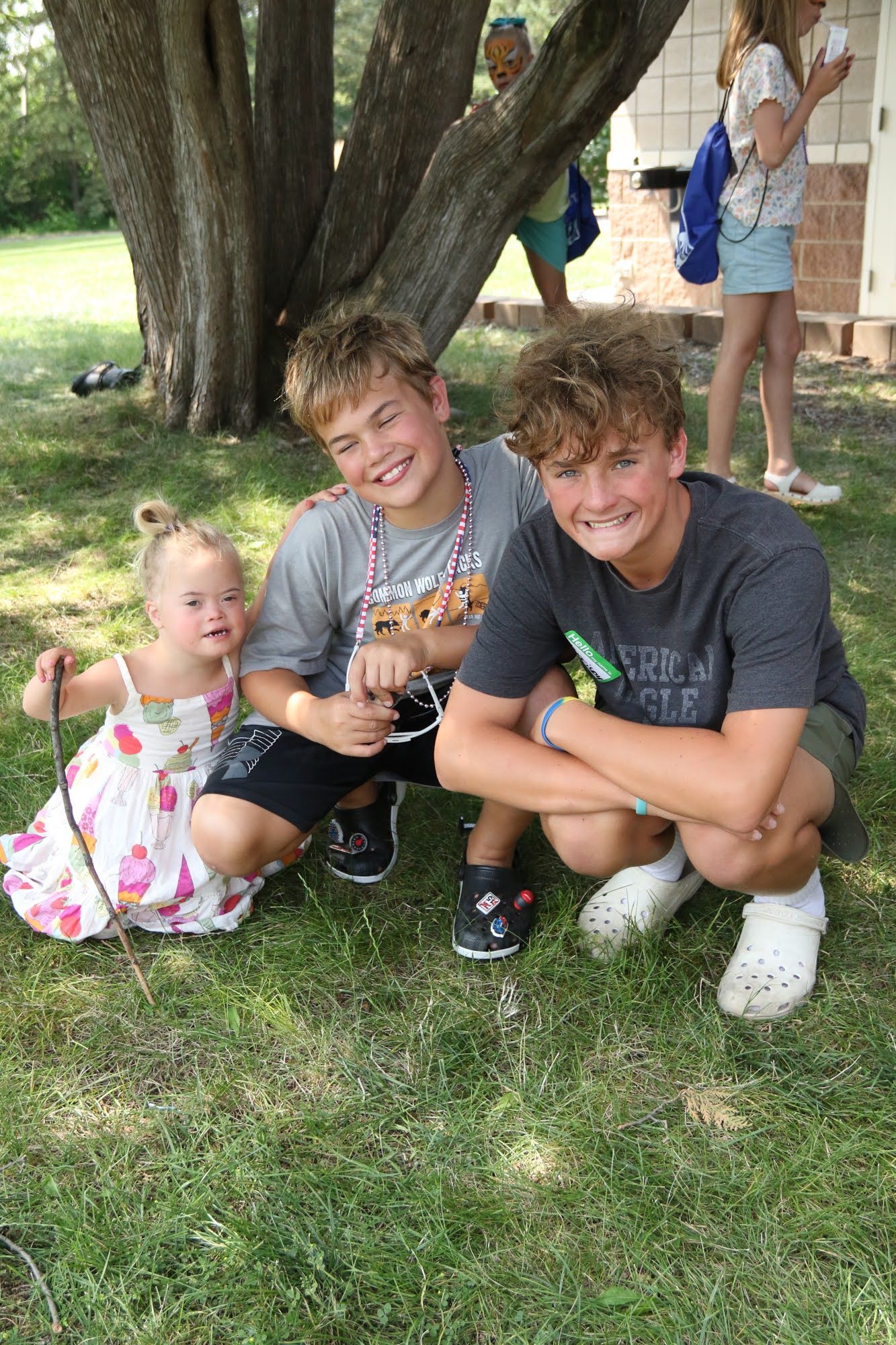 Three children squatting on grass, smiling. One in a dress, two in shirts and shorts. Outdoors near a tree.