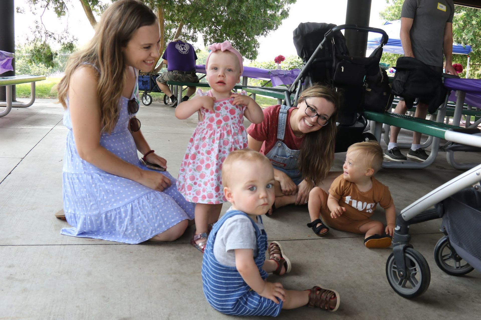 People and babies at outdoor event; children wear dresses and overalls, adults smile.
