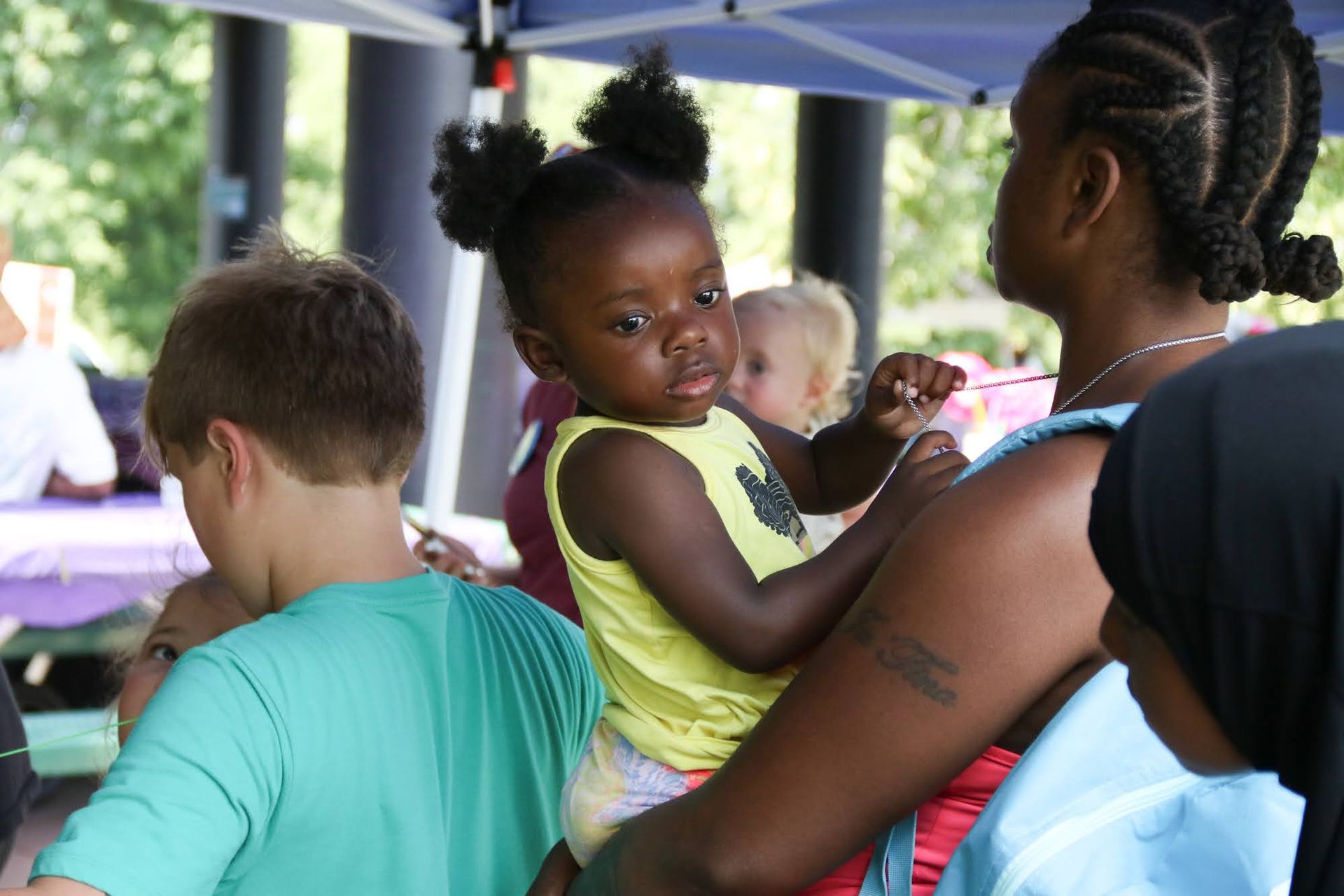 Child in arms at outdoor gathering, looking slightly to the side. Other people and a table are in the background.