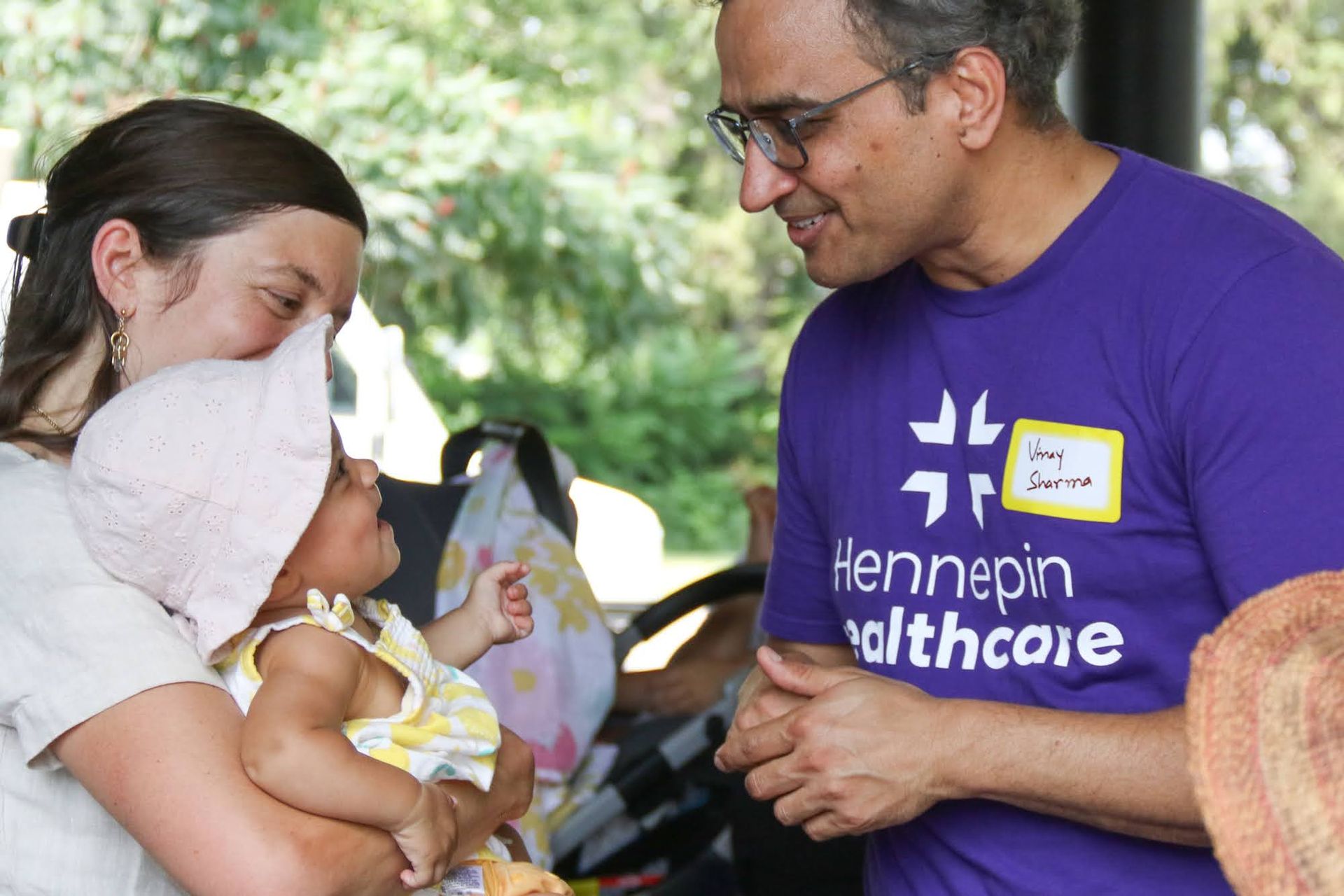 Man in purple shirt talks to a woman holding a baby. Outdoor setting.