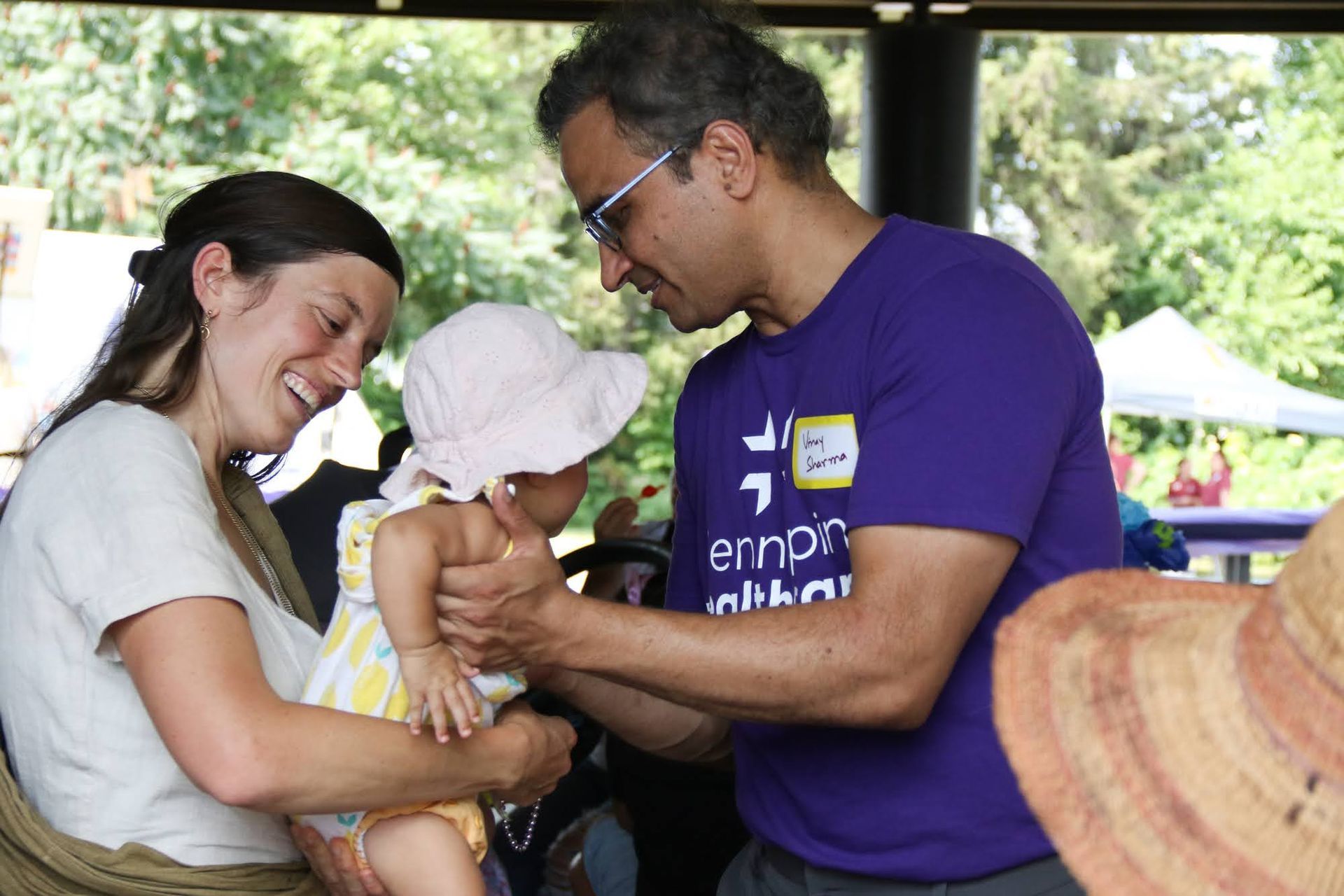 Woman holds toddler as man adjusts her hat; all are smiling. Outdoor setting.