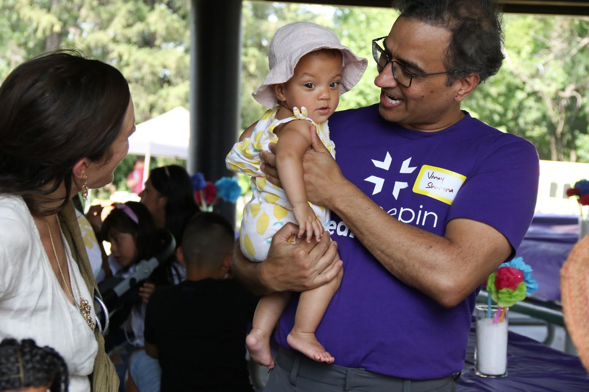 Man holding baby at outdoor event; woman looks on. Baby wears hat, man wears purple shirt.