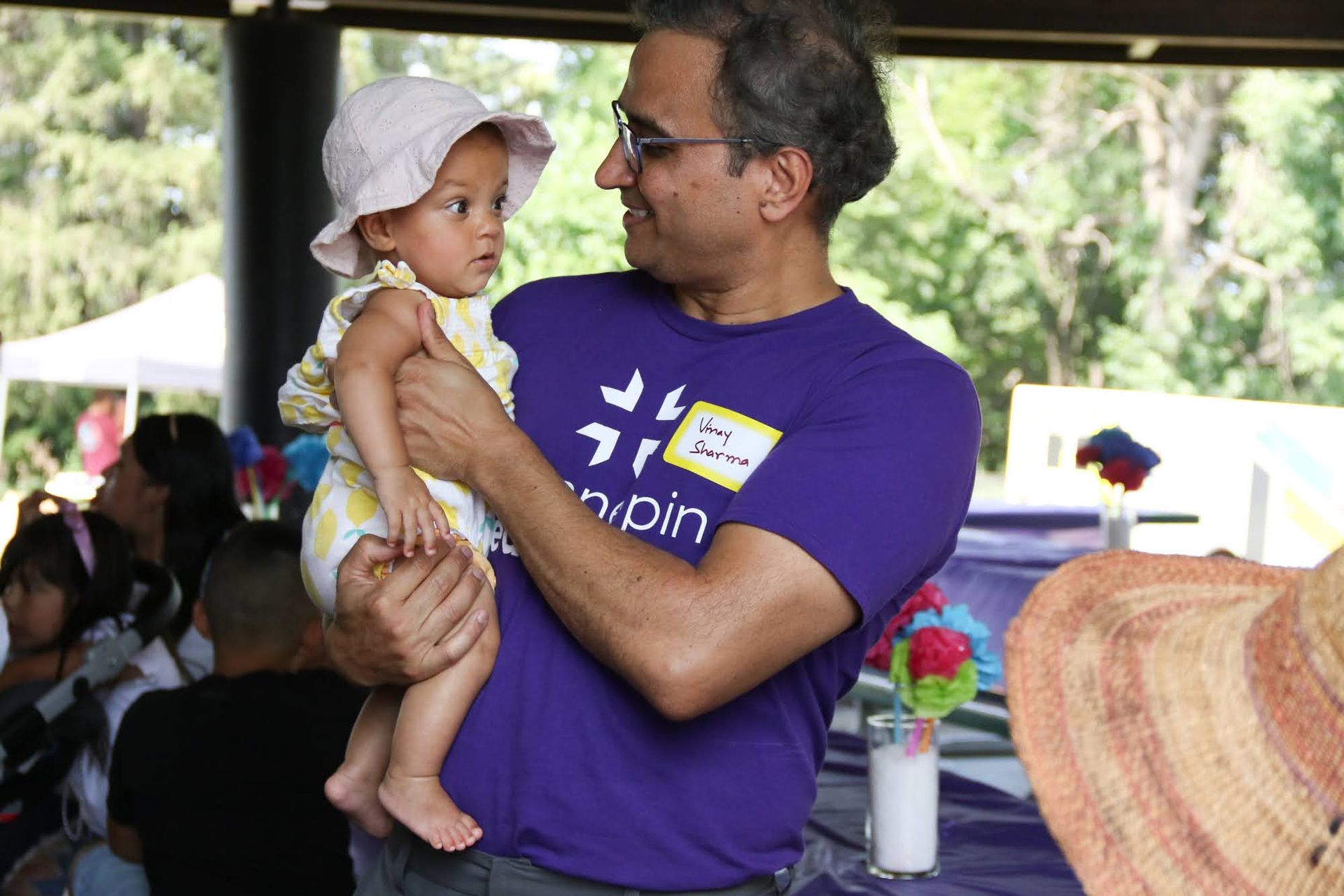 Man holding a baby, both looking at each other. Baby wears a hat and the man wears a purple shirt, possibly outdoors.