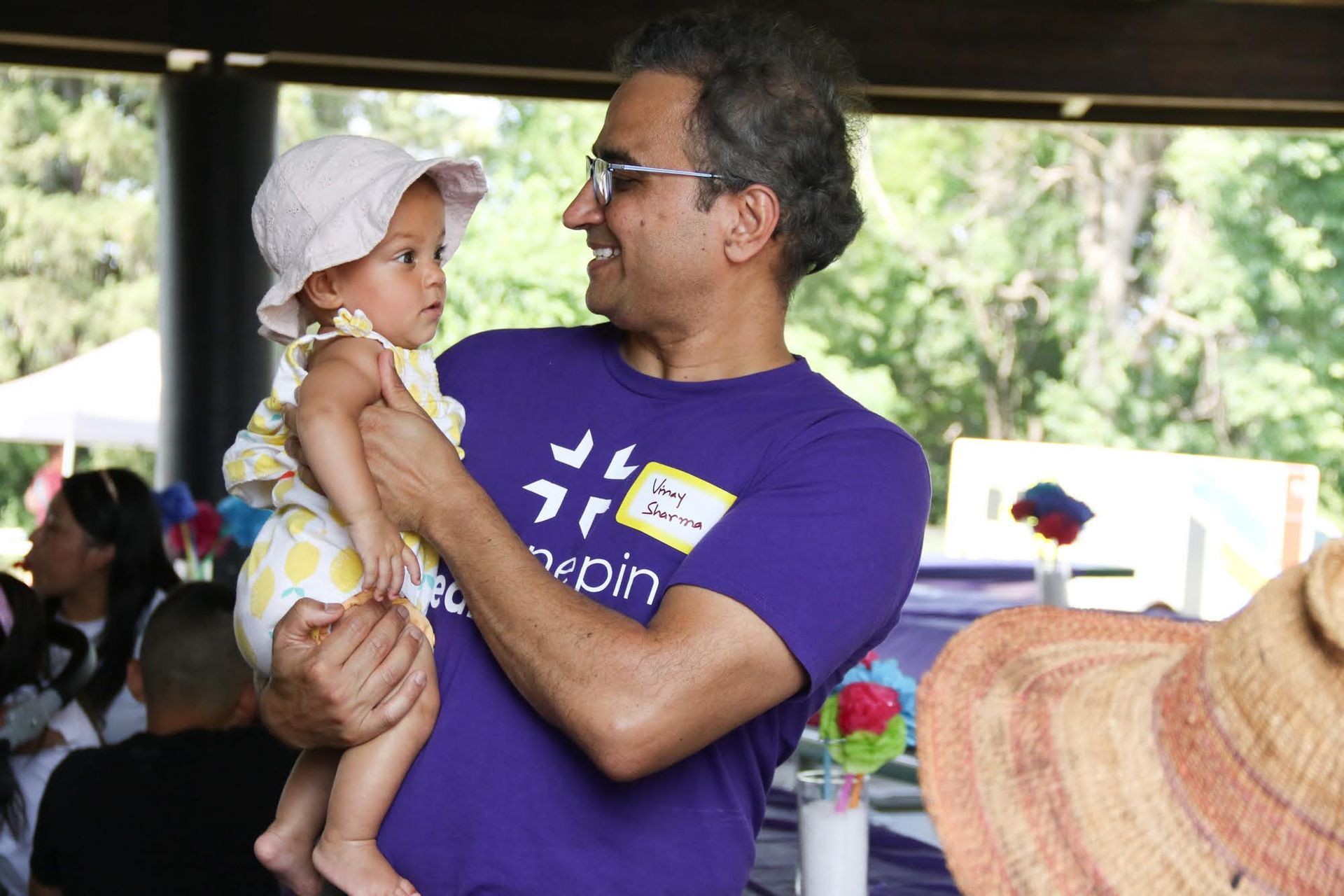 Man holding a baby wearing a hat outdoors, smiling at the child.  They are at a gathering with others in the background.