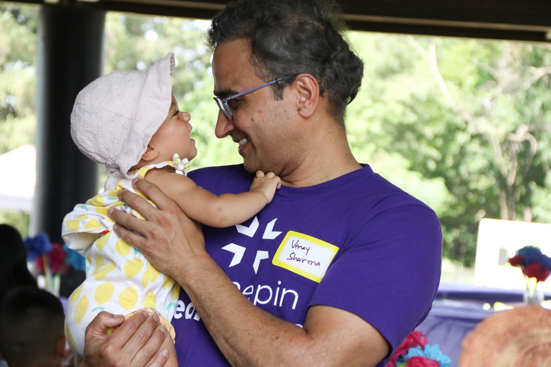 Man holding baby, both smiling at each other. Baby wears a hat and yellow polka dot outfit. They are outdoors.
