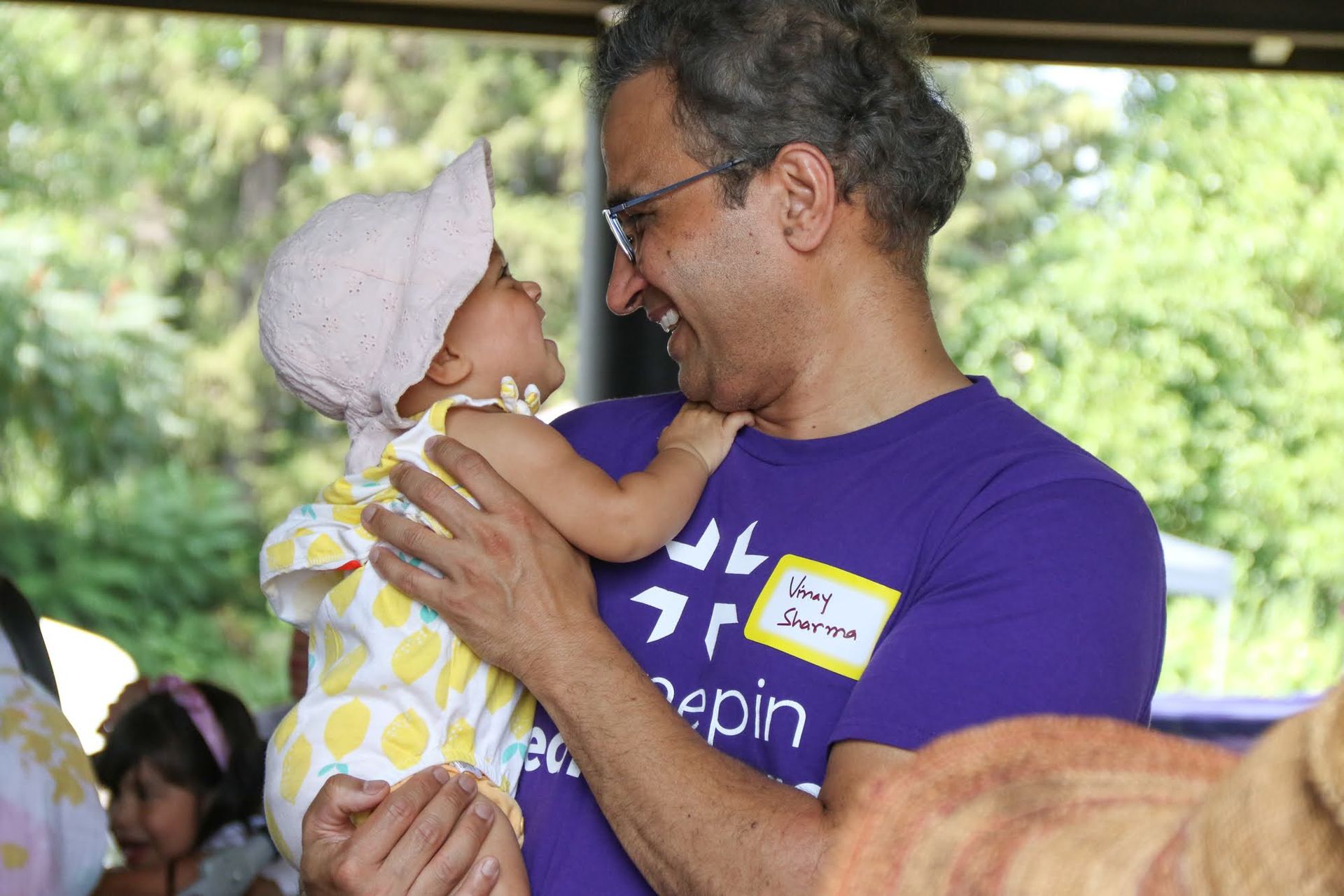 Man in purple shirt smiling, holding and looking at a baby in a sun hat, outdoors.