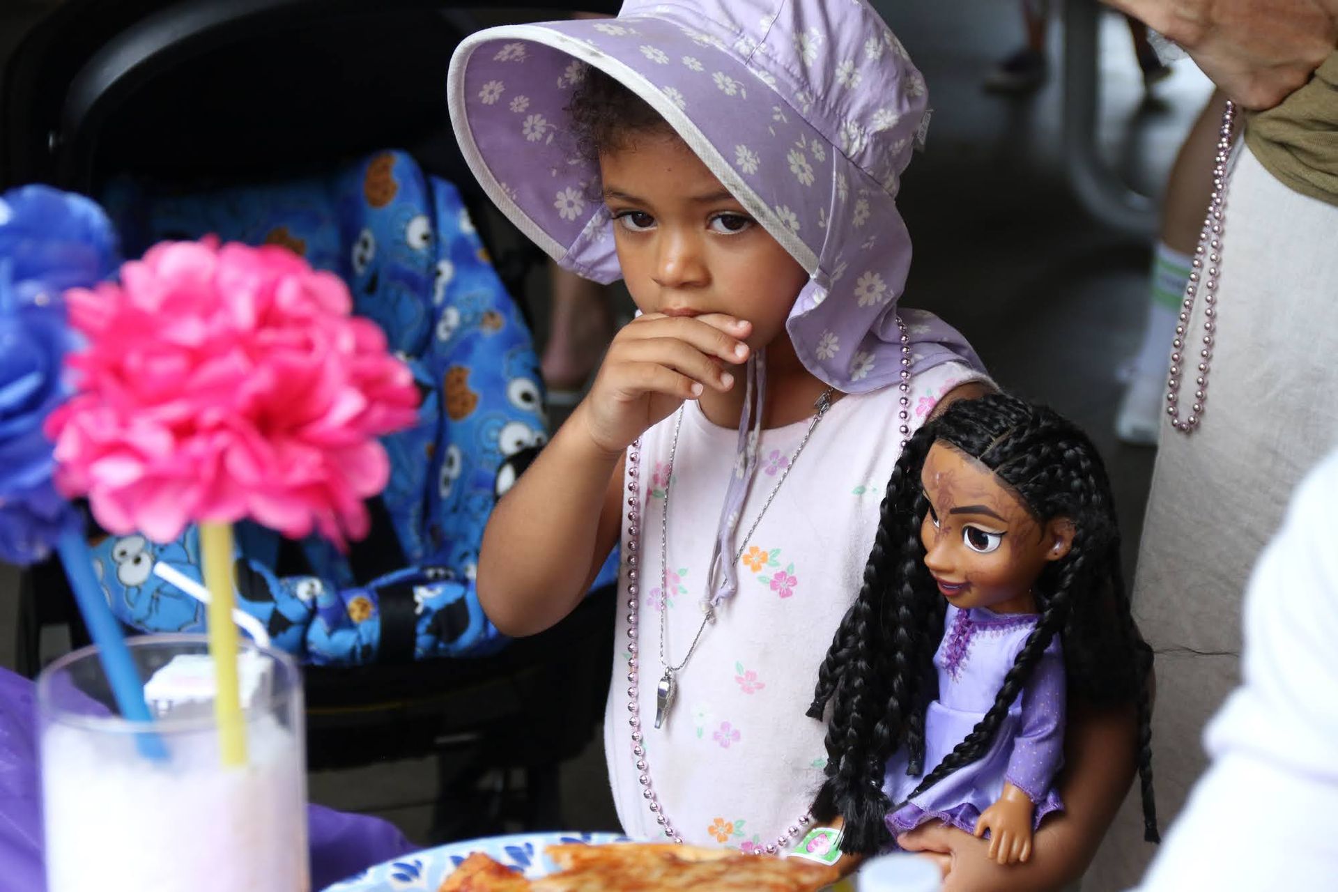 Girl in a sun hat holds a doll, looking thoughtful; a drink and flowers are nearby.