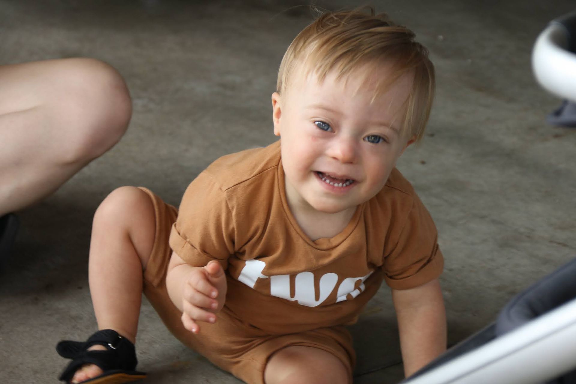 Smiling toddler in brown outfit with Down syndrome sits on floor.