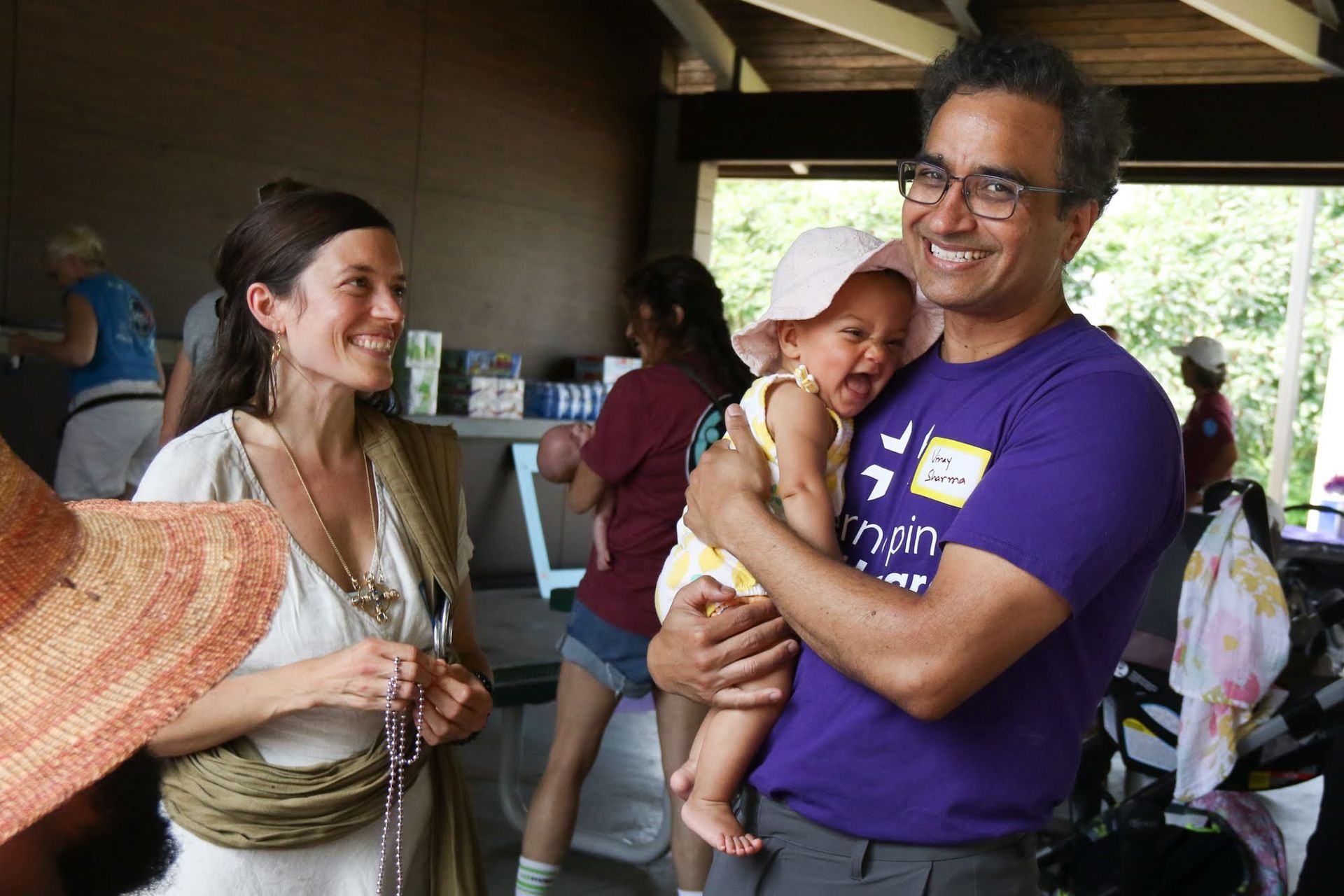 Man holding baby, smiling. Woman looks on. Outdoor gathering, people in background.