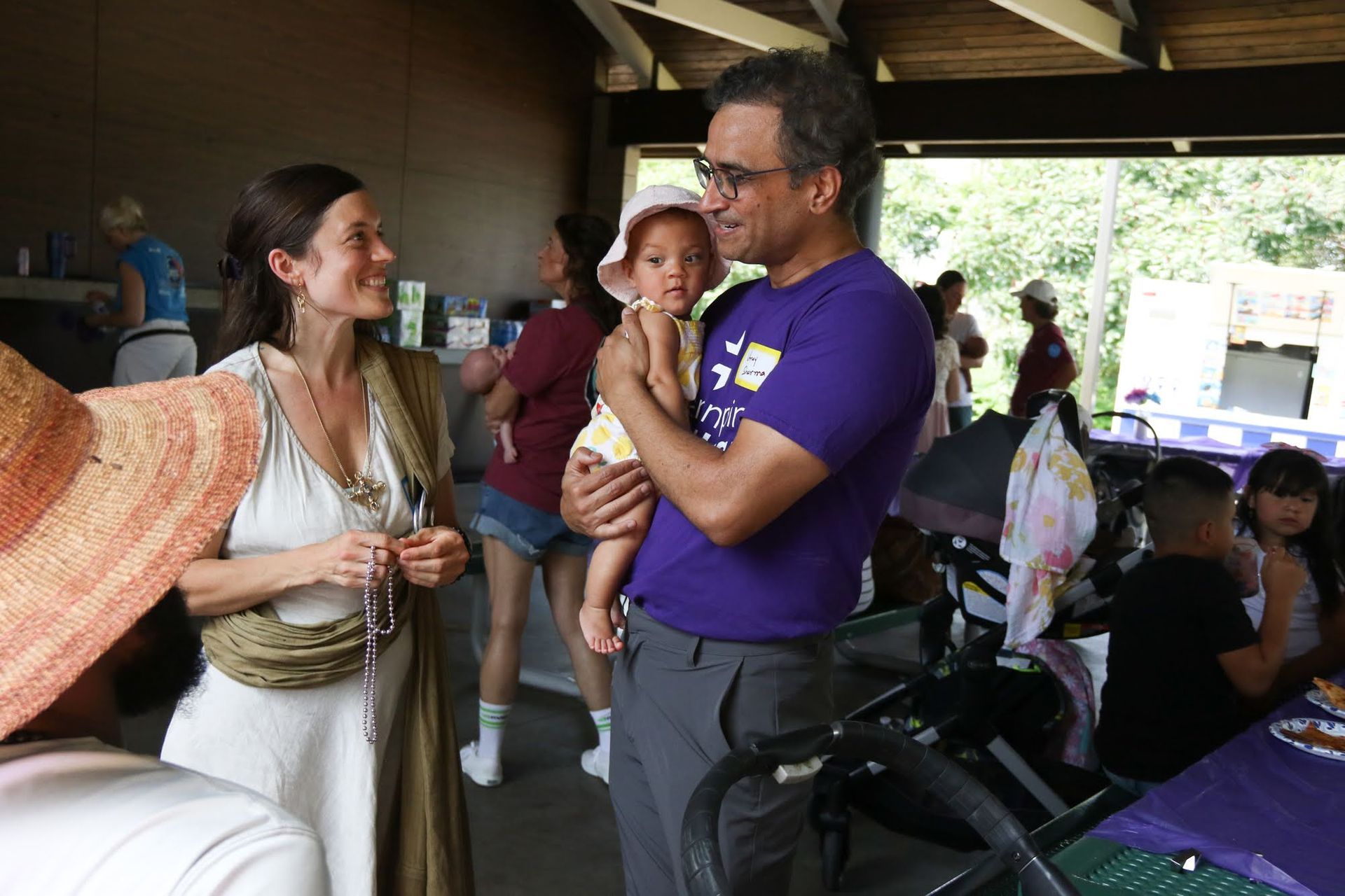 People at a park shelter. A man holds a baby; a woman smiles. Tables, other people are visible.