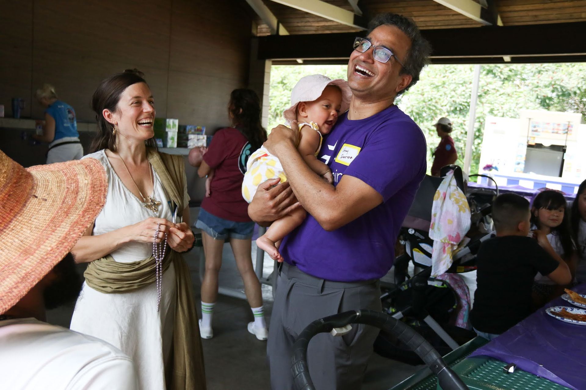 Man holding baby laughs, woman smiles at family gathering in park pavilion.