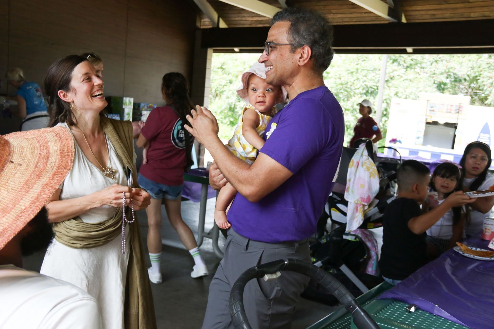 Man holding baby smiles at woman, at outdoor gathering.