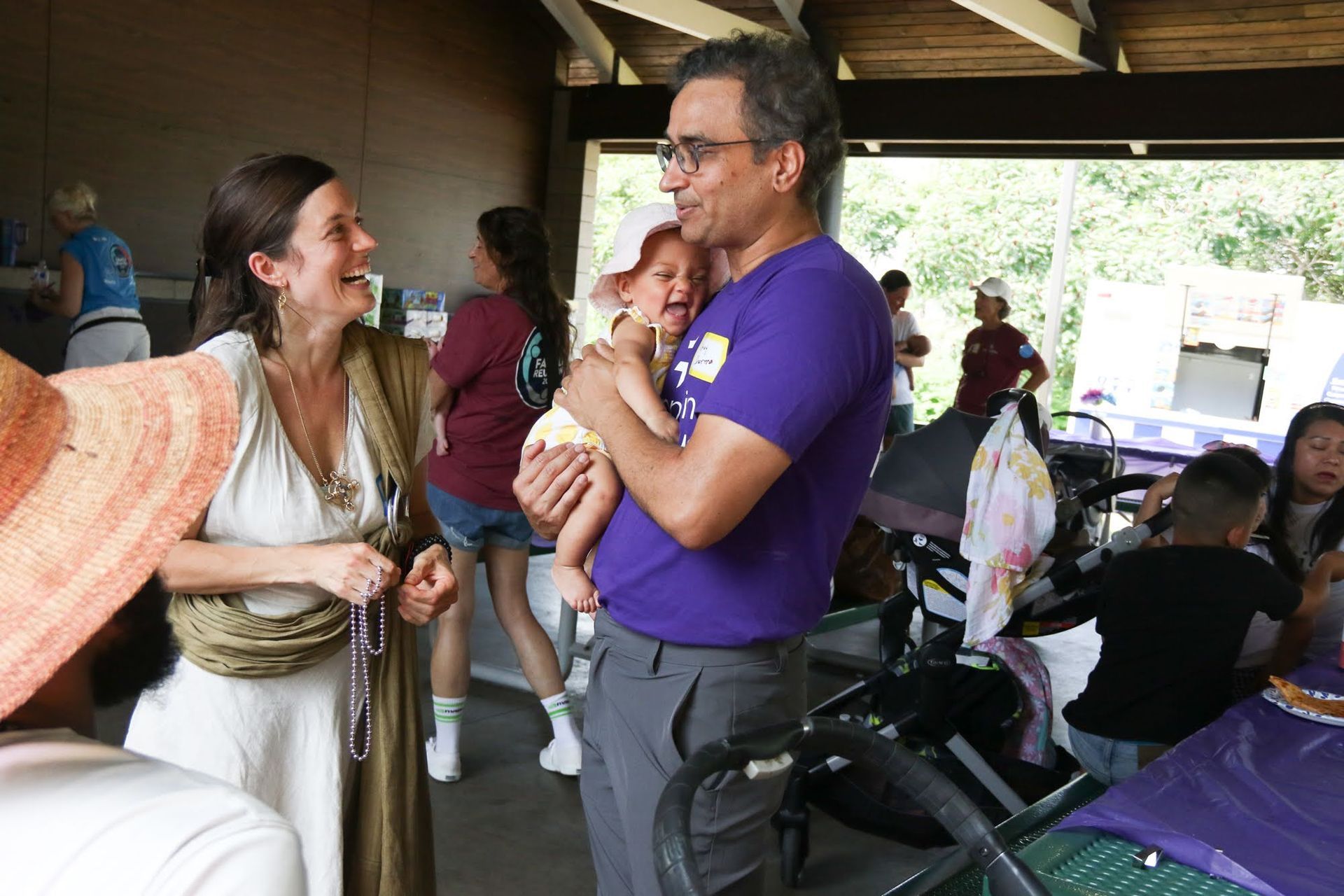 Woman and man talking, man holding baby in a park shelter, other people in the background.