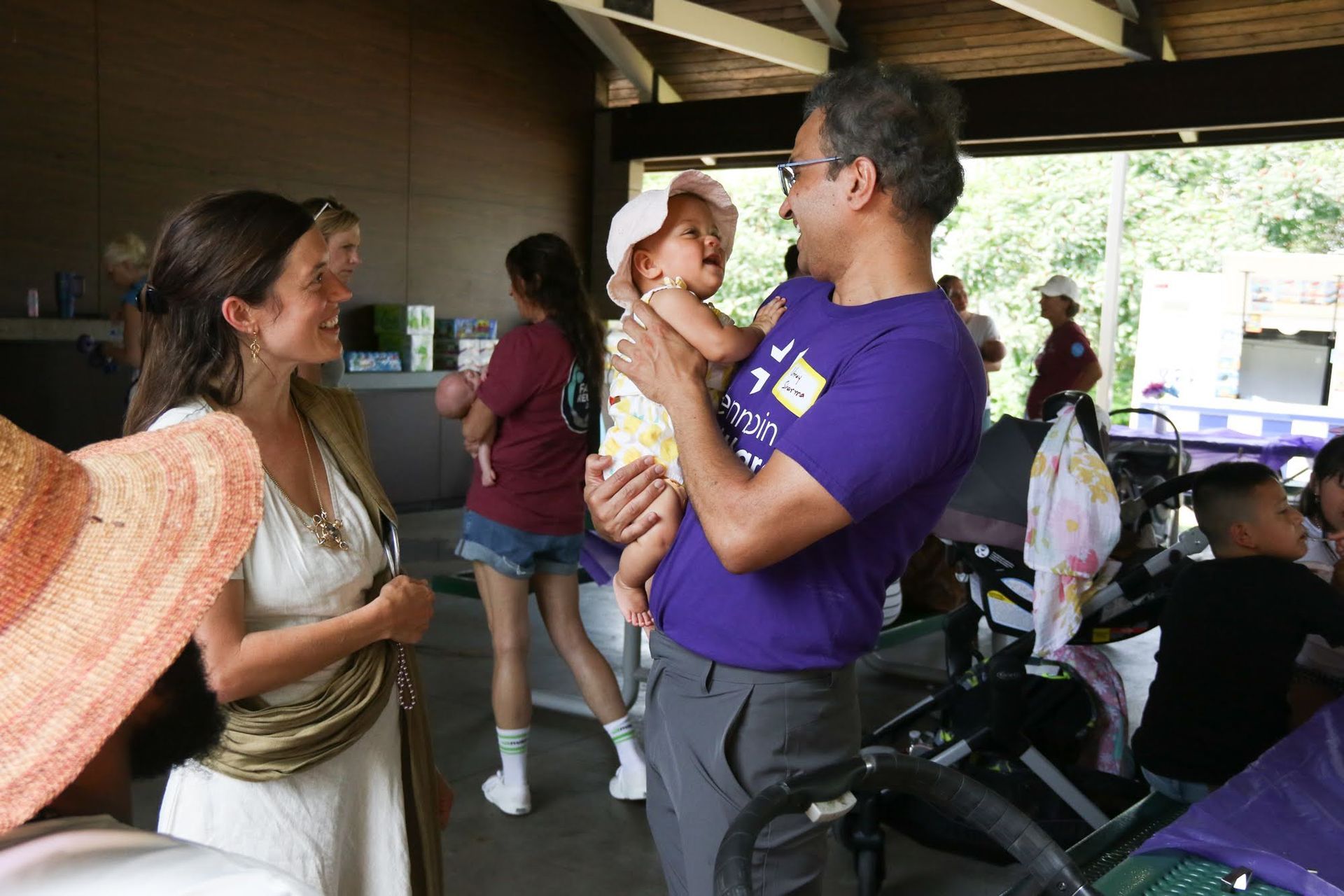 Man holding baby smiles at woman, other people in background at outdoor gathering.