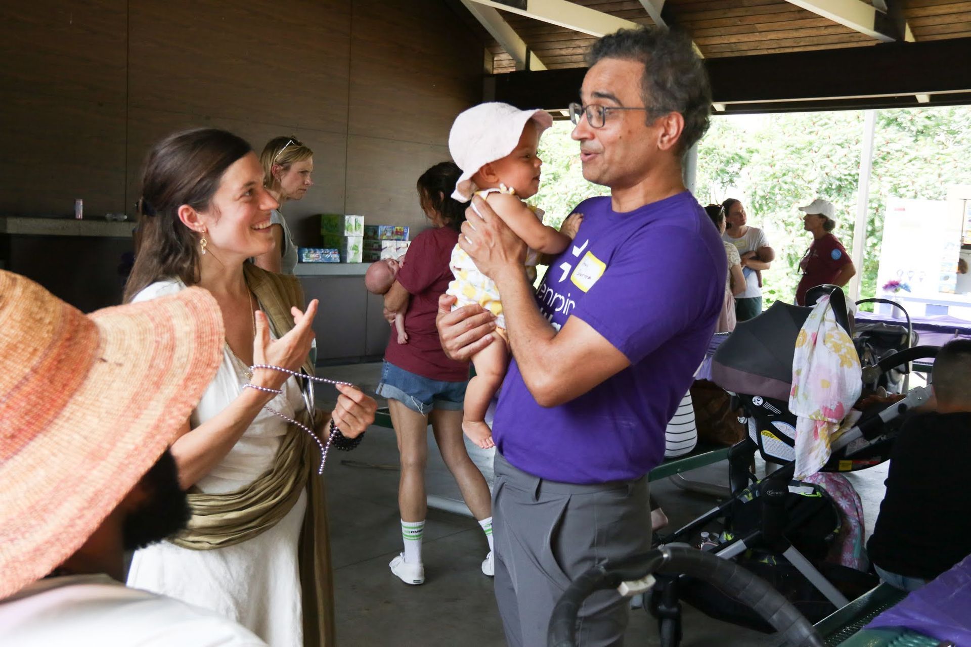 Man in purple shirt holds baby, talking with woman in white dress at outdoor gathering.